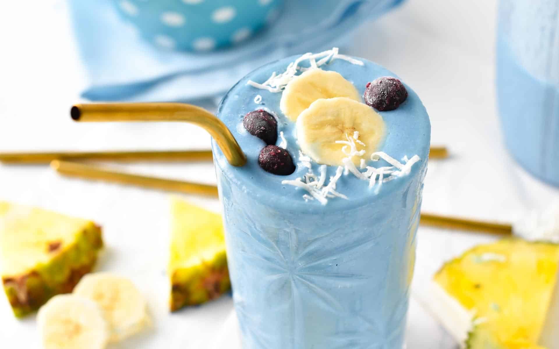 Blue spirulina smoothie glass decorated with a slice of banana, blueberries, and shredded coconut with a golden straw.