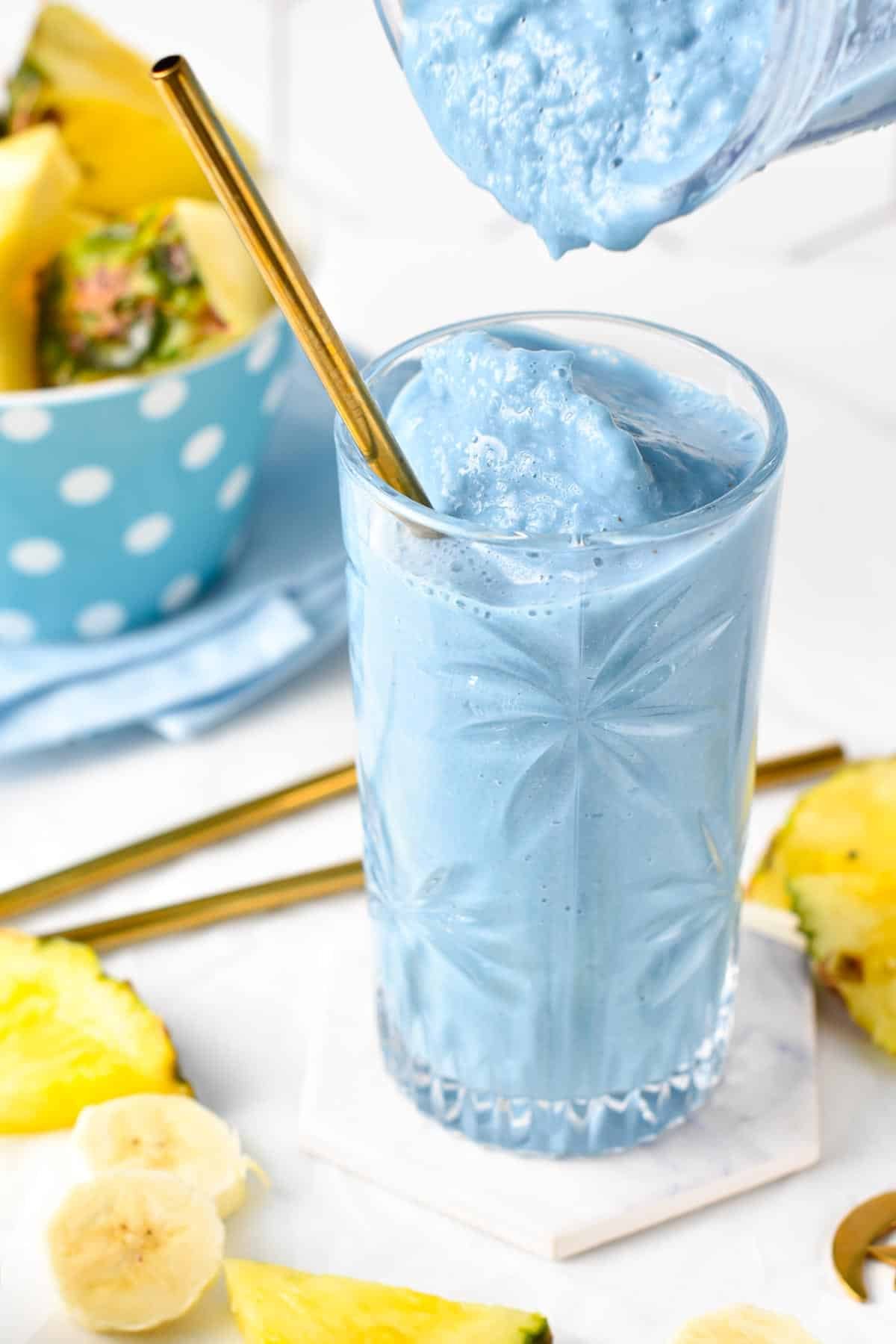 Pouring blue spirulina smoothie into a tall glass with a golden straw.