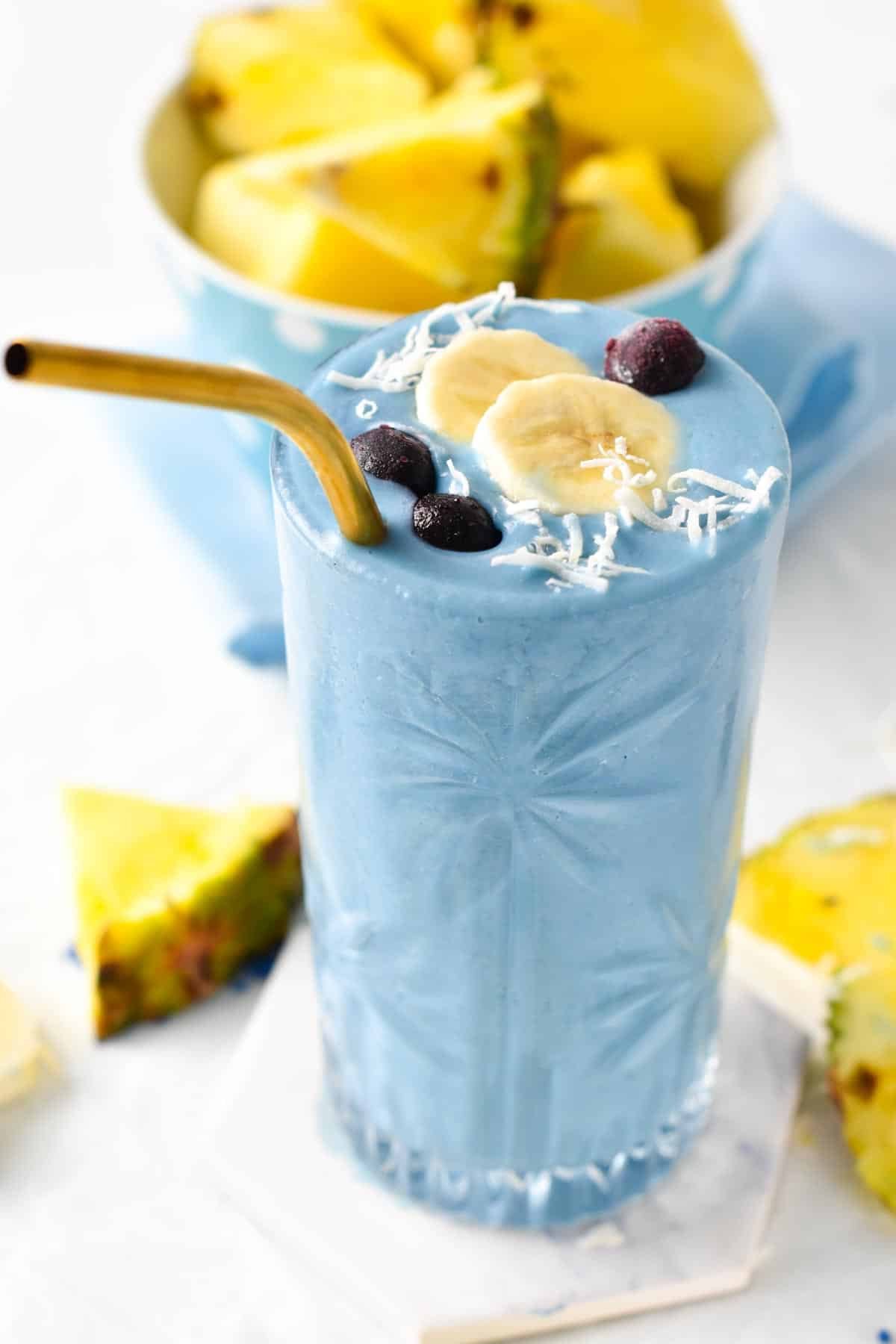 Glass full of blue spirulina smoothie decorated with a slice of banana, blueberries, and shredded coconut with a golden straw.