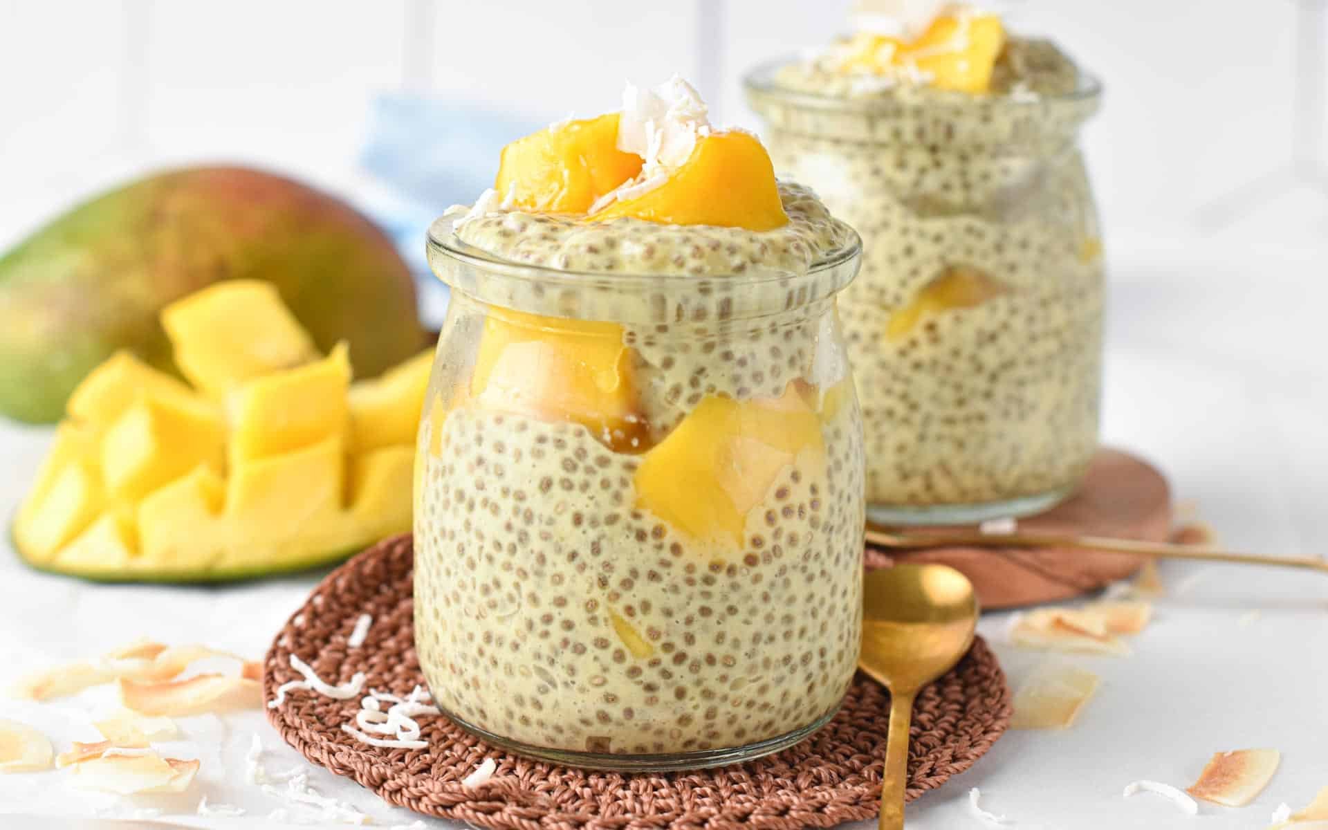 Mango chia pudding in two small jars on brown coasters with a golden spoon and a cut mango in the background.