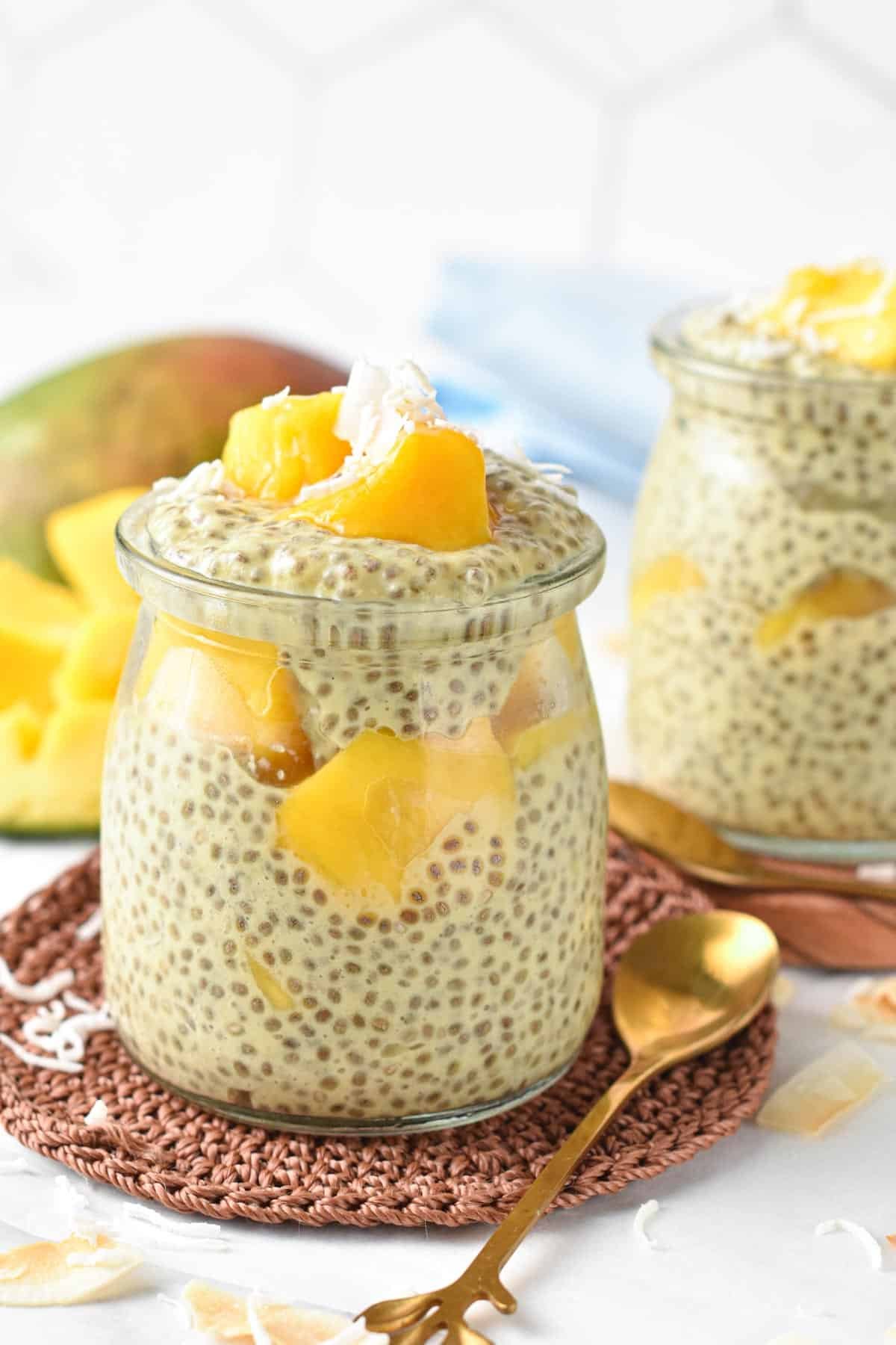 Mango chia pudding in two small jars on brown coasters with a golden spoon.