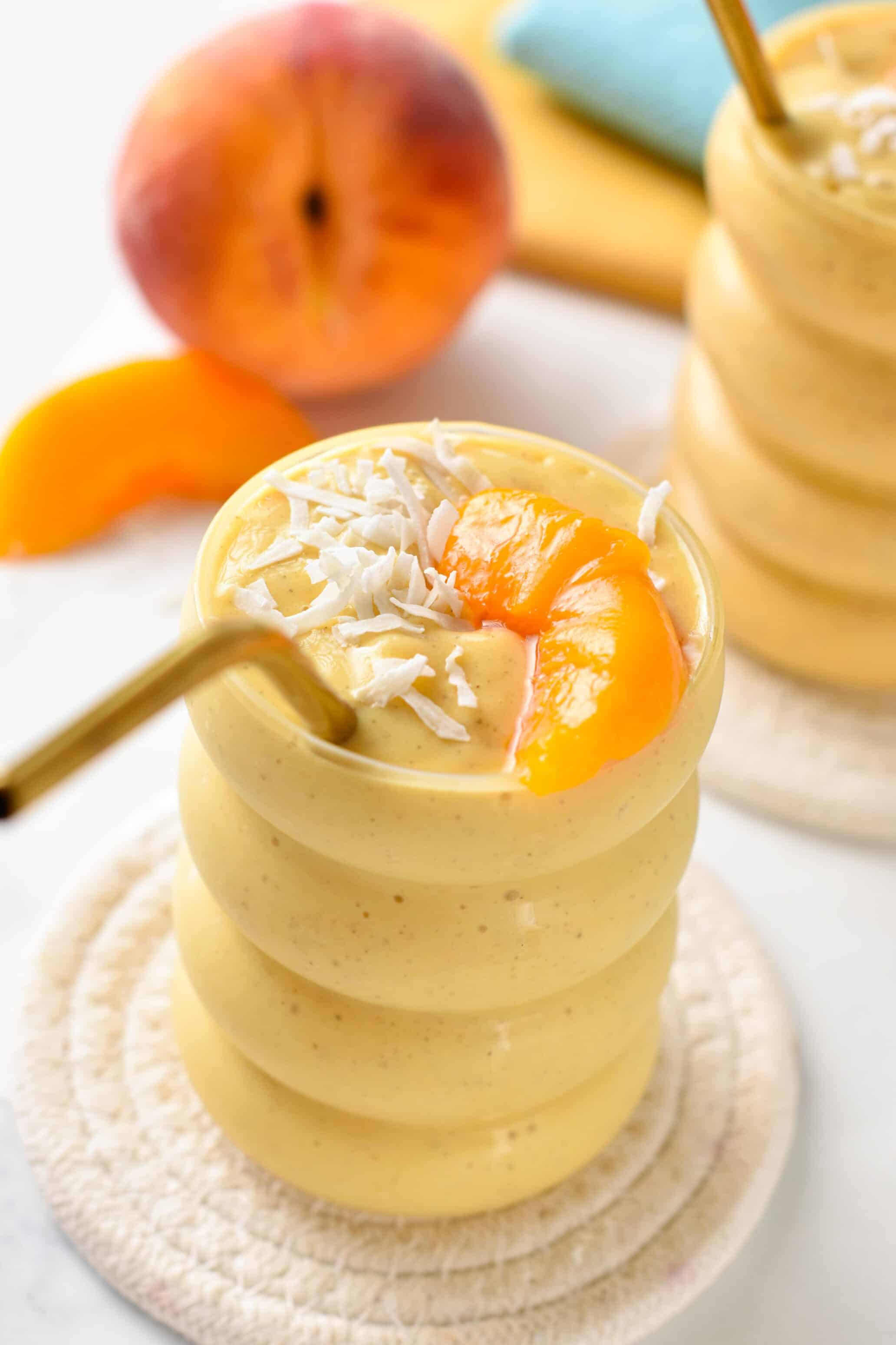 Banana Peach Smoothie in a tall glass with a golden straw, decorated with fresh peach and shredded coconut.