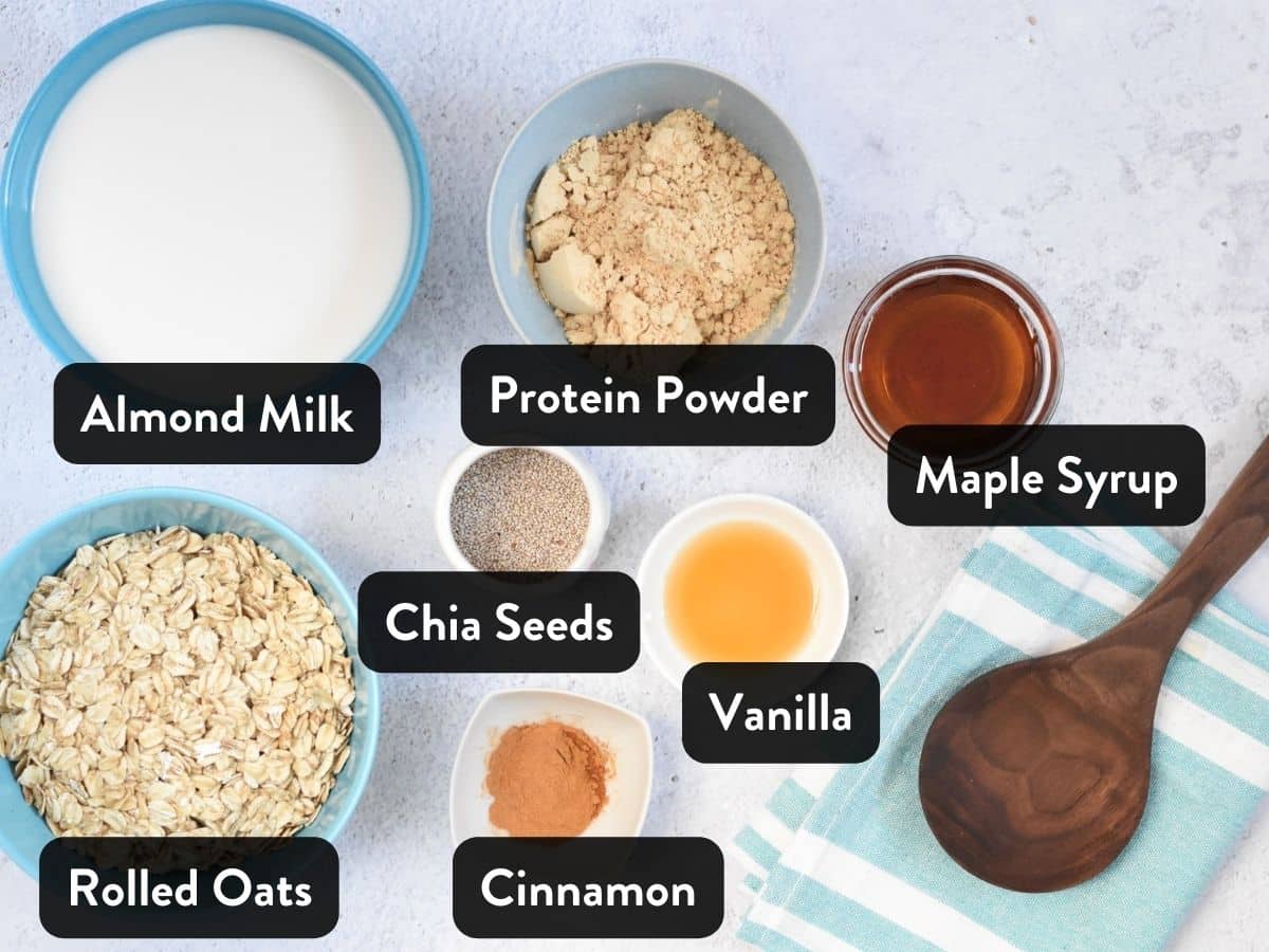 Ingredients for Overnight Oats with Protein Powder organized in various bowls on a kitchen benchtop next to a wooden spoon.