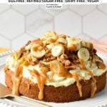 a single layer oat flour cake topped with dairy-free vanilal frosting, banana slices, walnuts, pecans, and peanut butter caramel