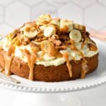 a single layer oat flour cake topped with dairy-free vanilal frosting, banana slices, walnuts, pecans, and peanut butter caramel