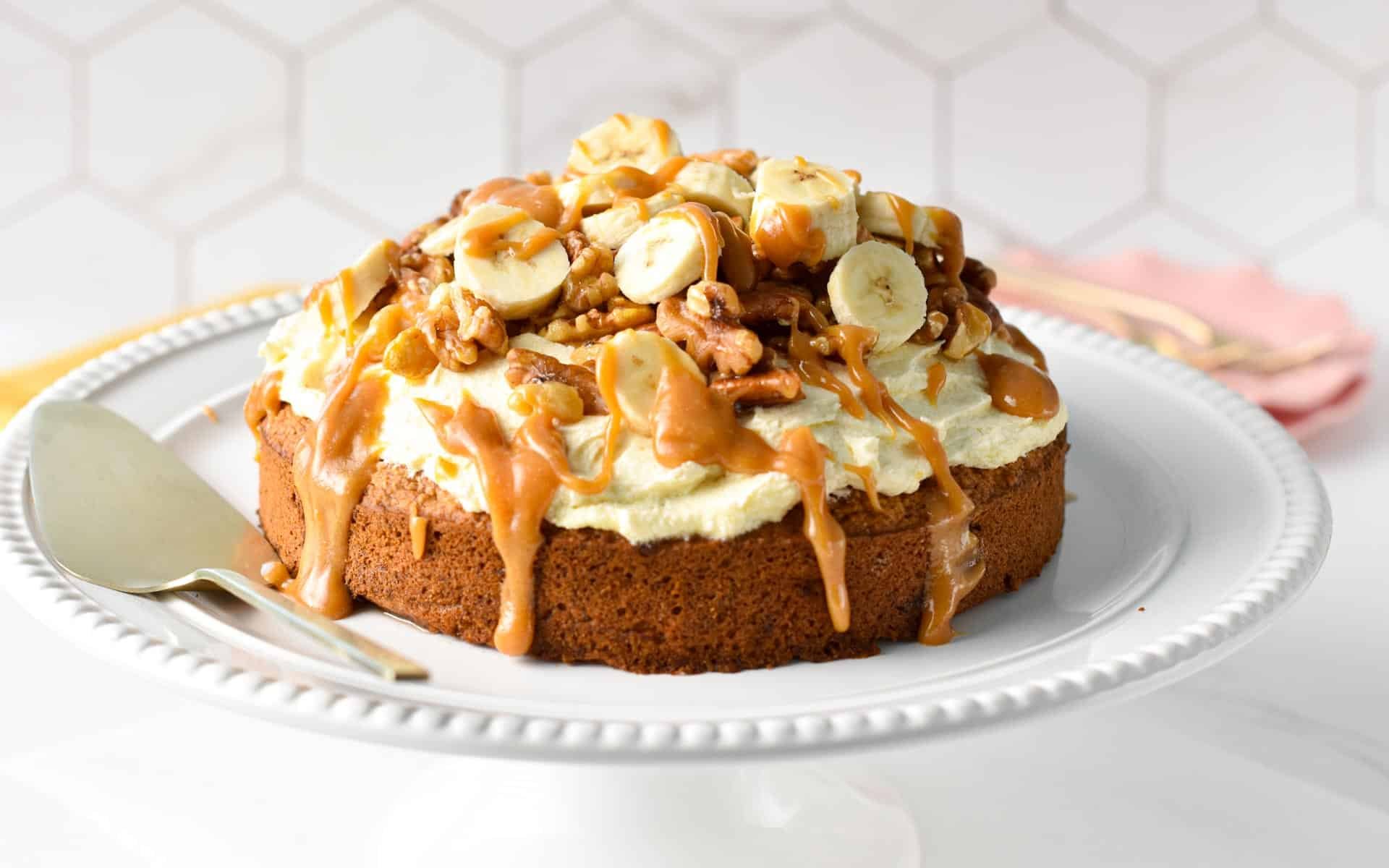 A single layer oat flour cake topped with dairy-free vanilla frosting, banana slices, walnuts, pecans, and peanut butter caramel on a cake stand with a serving spatula next to it.