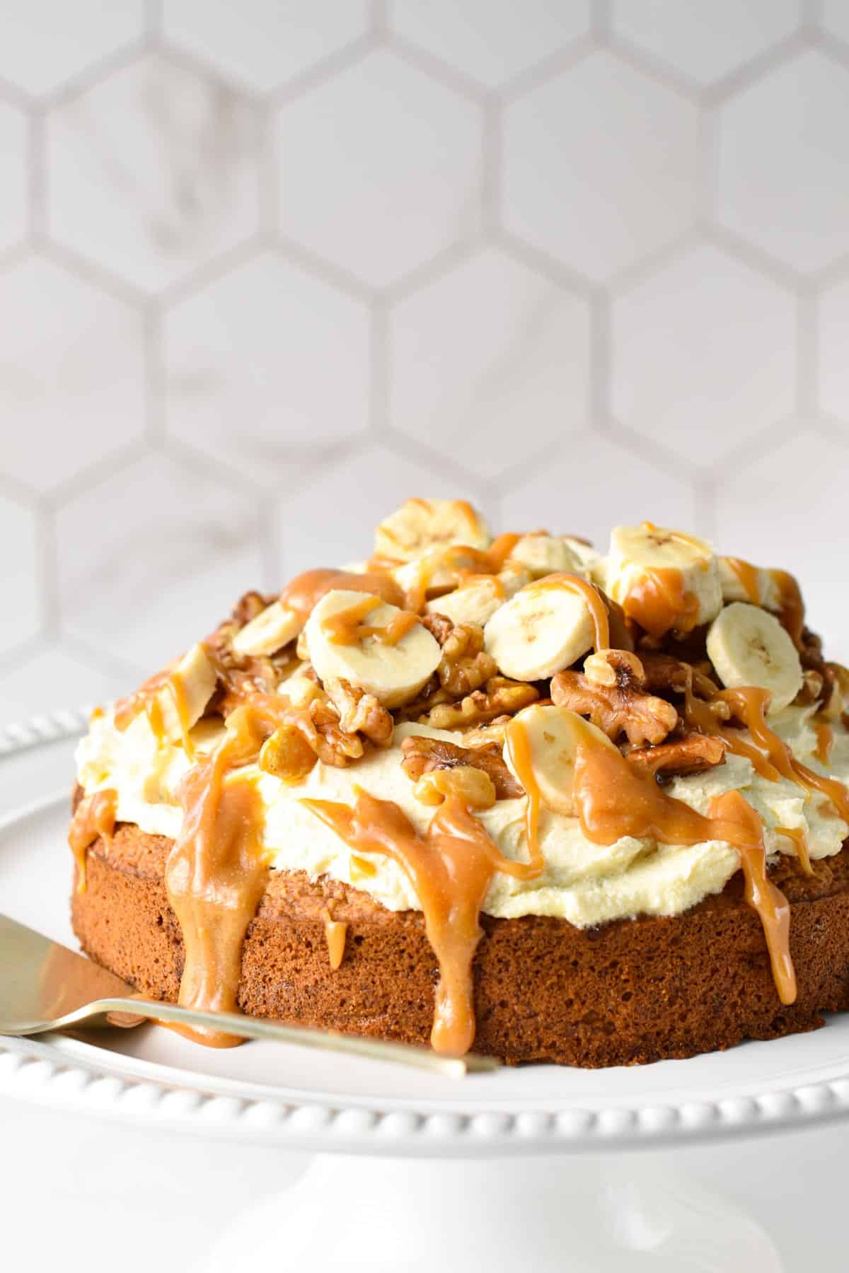 A single layer oat flour cake topped with dairy-free vanilla frosting, banana slices, walnuts, pecans, and peanut butter caramel.