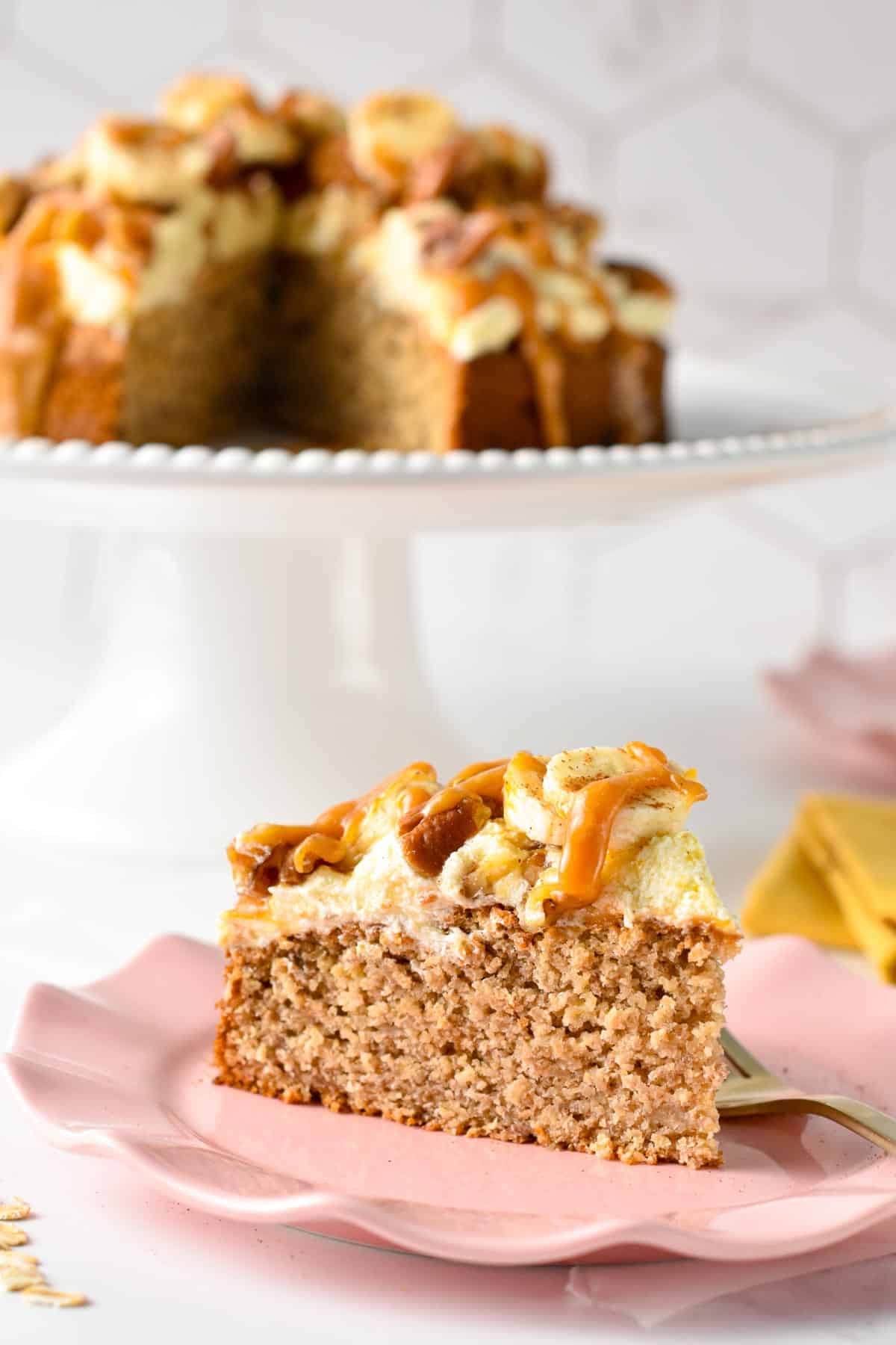 A slice of single layer oat flour cake topped with dairy-free vanilla frosting, banana slices, walnuts, pecans, and peanut butter caramel on a small pink plate with golden forks