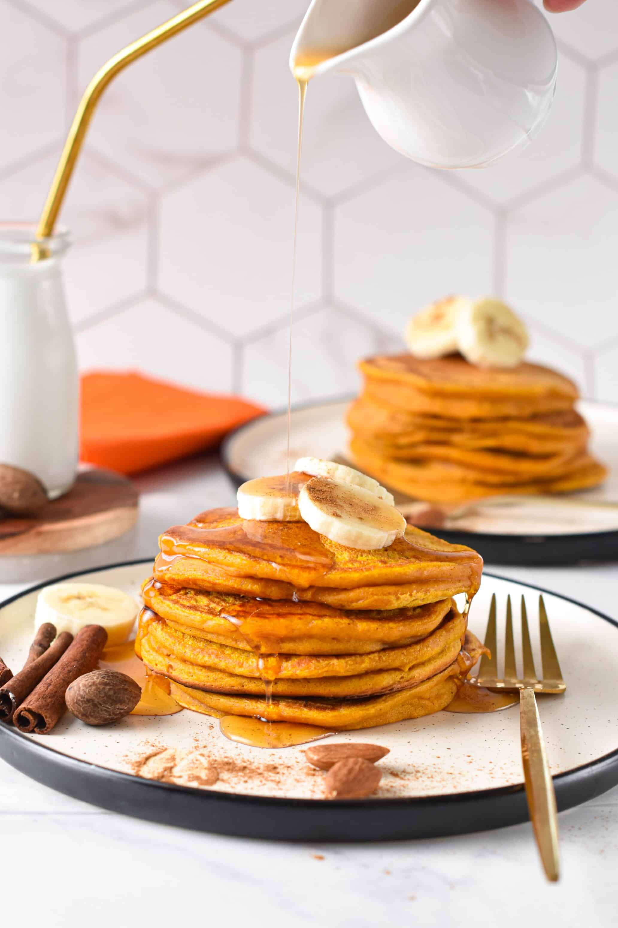 Pumpkin Protein Pancakes stacked on a plate with banana slices, cinnamon sticks, and a drizzle of maple syrup.