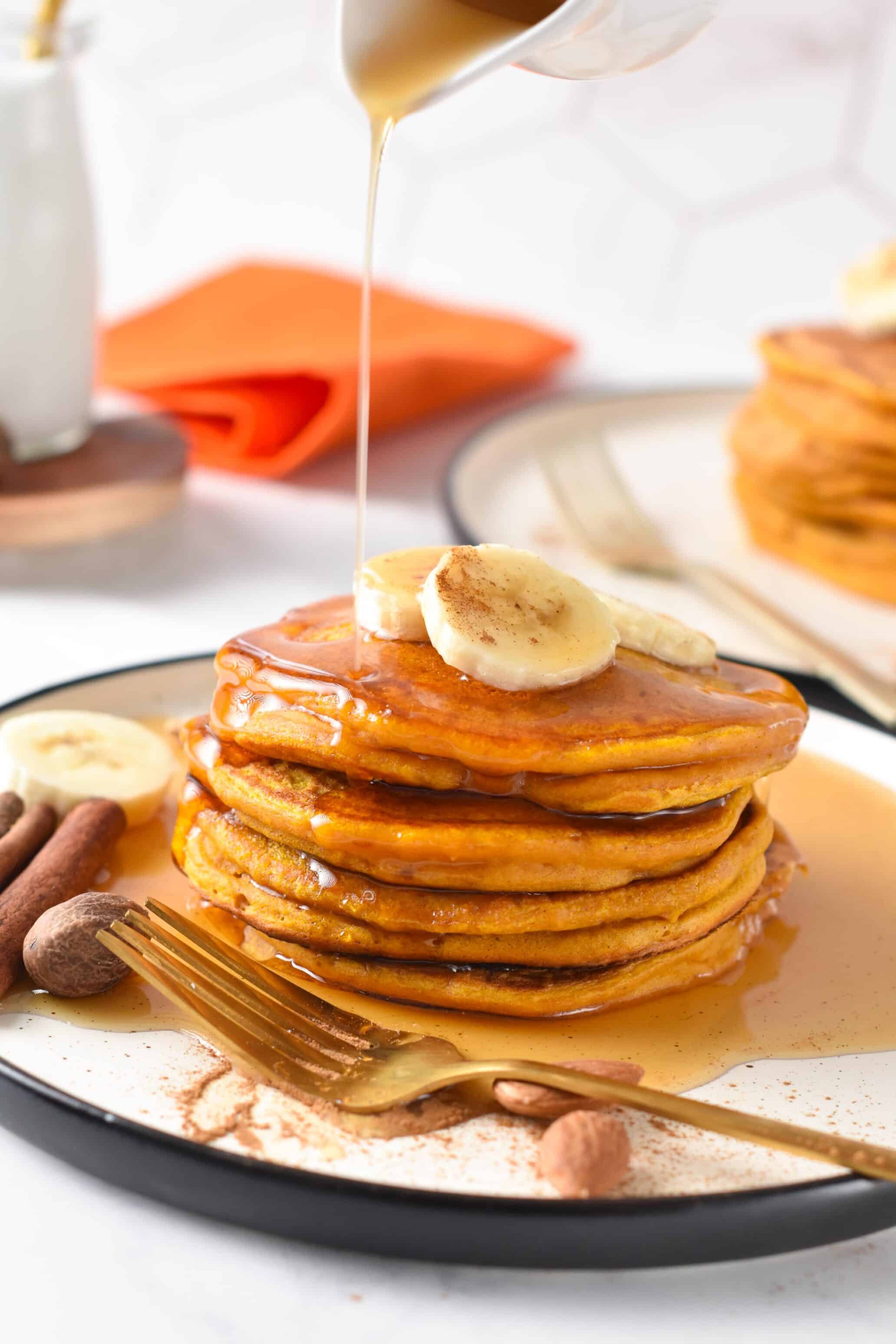 Stack of Pumpkin Protein Pancakes on a plate with banana slices and maple syrup drizzling on them