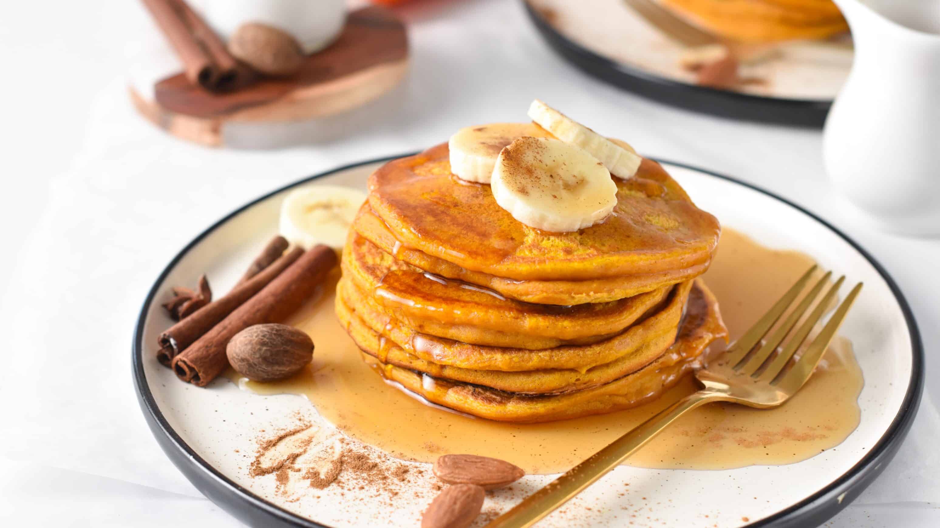 Stack of Pumpkin Protein Pancakes on a plate with banana slices, cinnamon sticks, almonds, and a golden fork.