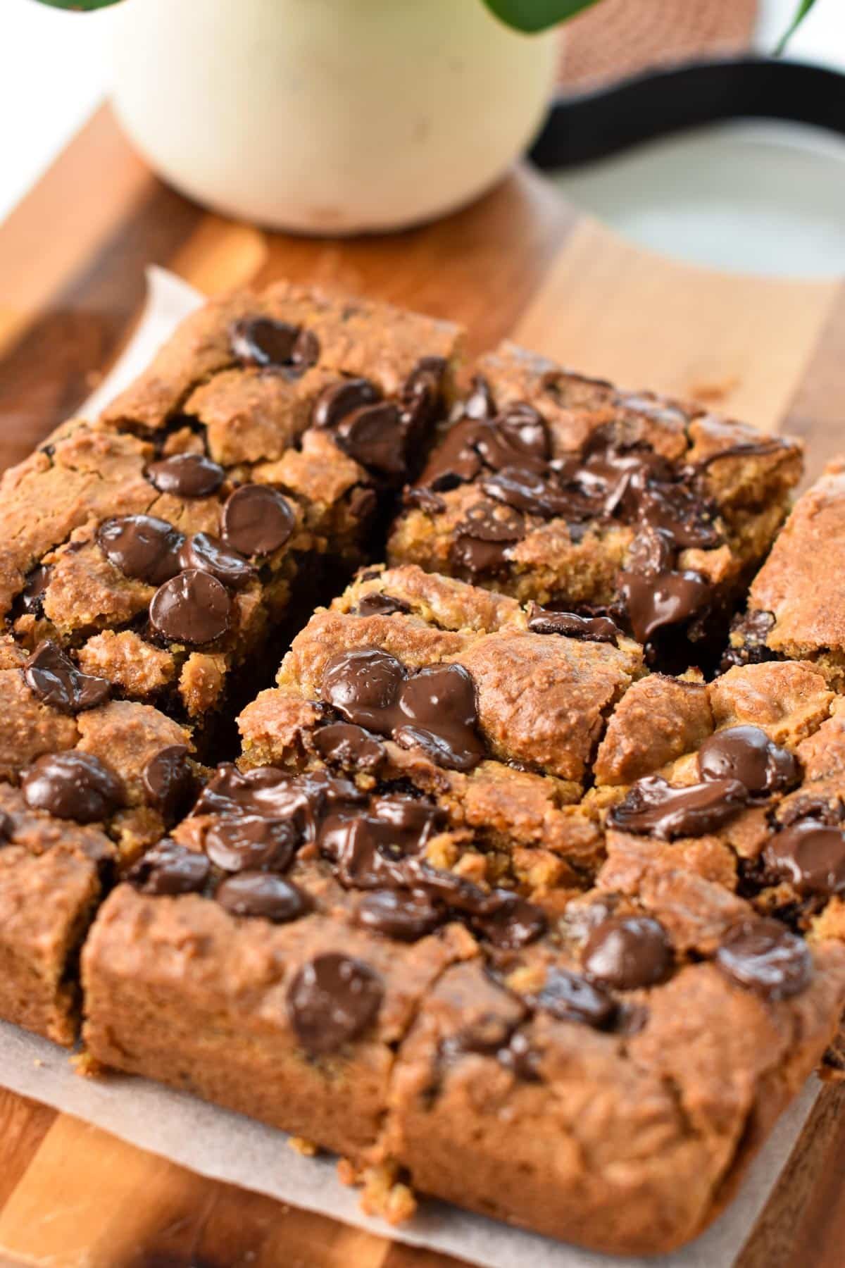 A wooden chopping board with lots of square of chickpea blondies with melted chocolate chips on top.