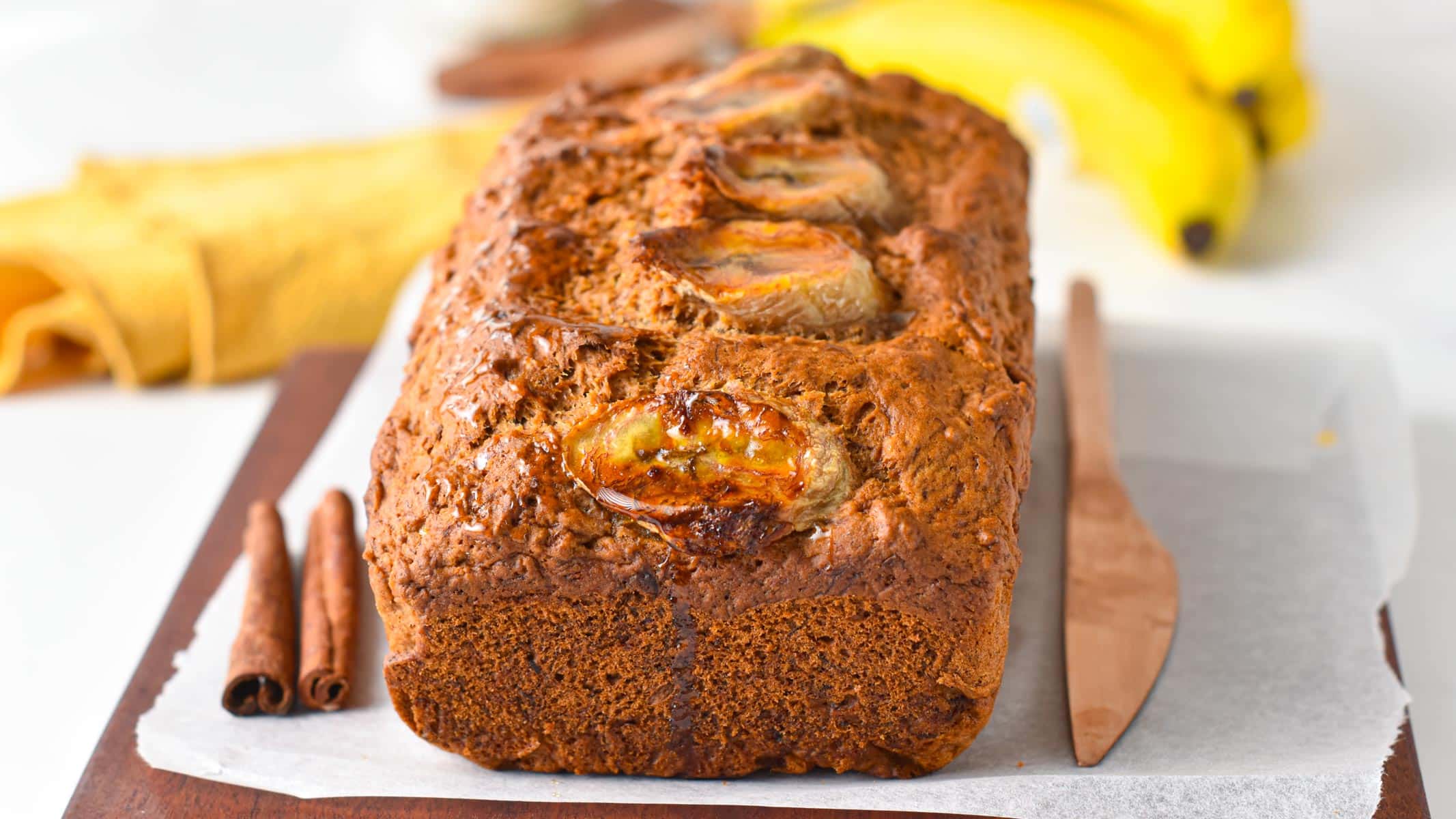 A banana bread made with spelt flour and banana slices on top on a chopping board with cinnamon sticks and a bronze knife.
