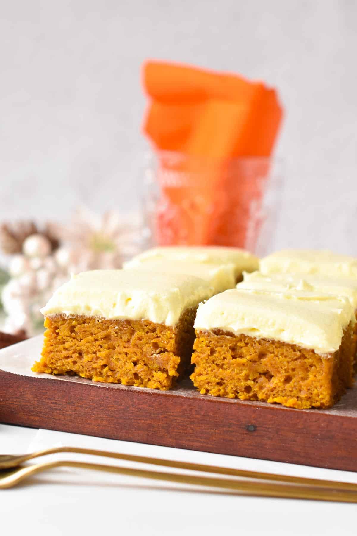 Slices of vegan pumpkin bars frosted with dairy-free vanilla frosting on a wooden chopping board behind two golden forks.