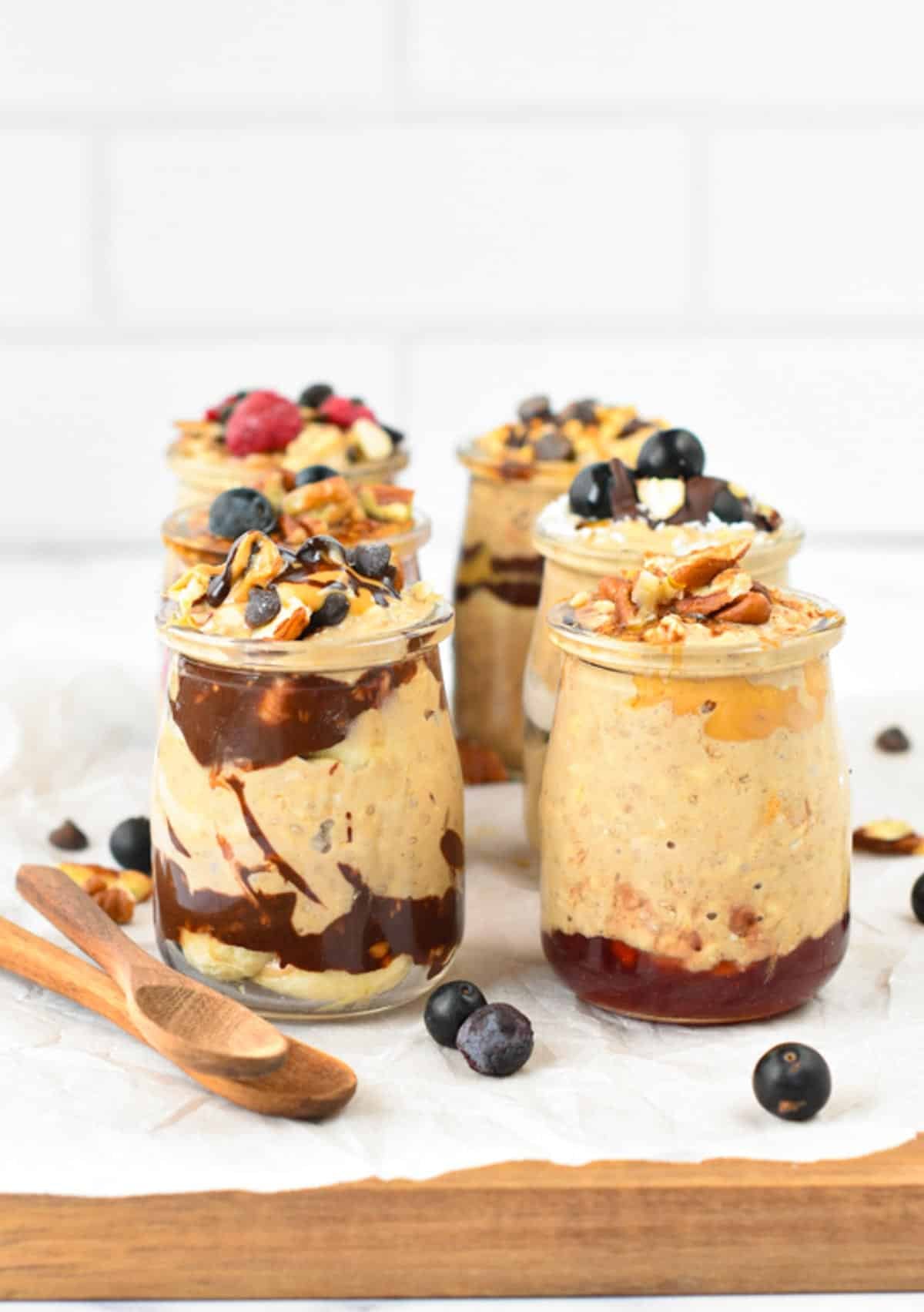 Small glass mason jars filled with protein overnight oats, and layers of different ingredients like berries, melted chocolate, peanut butter, and nuts.