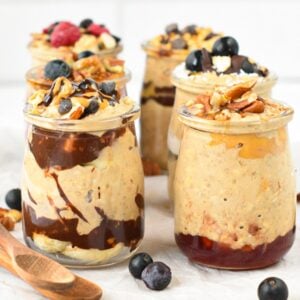 Small glass mason jars filled with protein overnight oats, and layers of different ingredients like berries, melted chocolate, peanut butter, and nuts