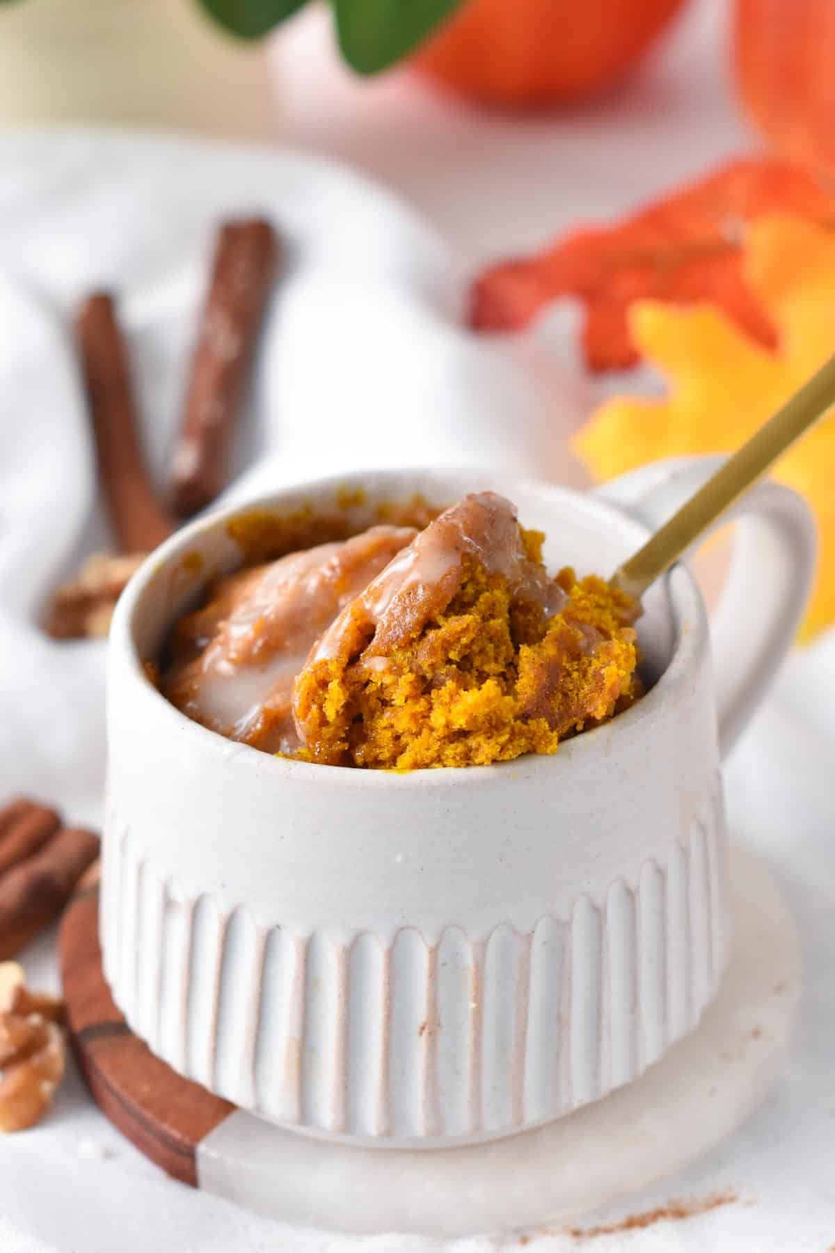 A mug filled with a pumpkin spice mug cake topped with a drizzle of vegan royal icing and a golden spoon digging in the mug cake.