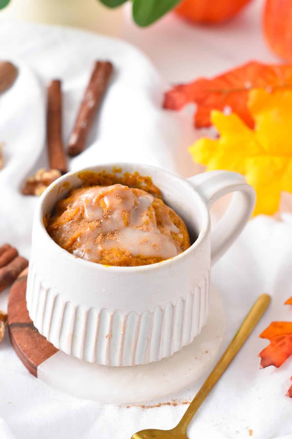 A mug filled with a pumpkin mug cake topped with a drizzle of vegan royal icing next to a golden spoon.