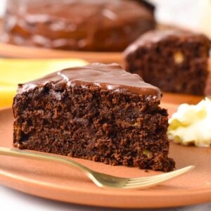 a slice of vegan chocolate banana cake with chocolate ganache, on a brown plate with golden fork in the front