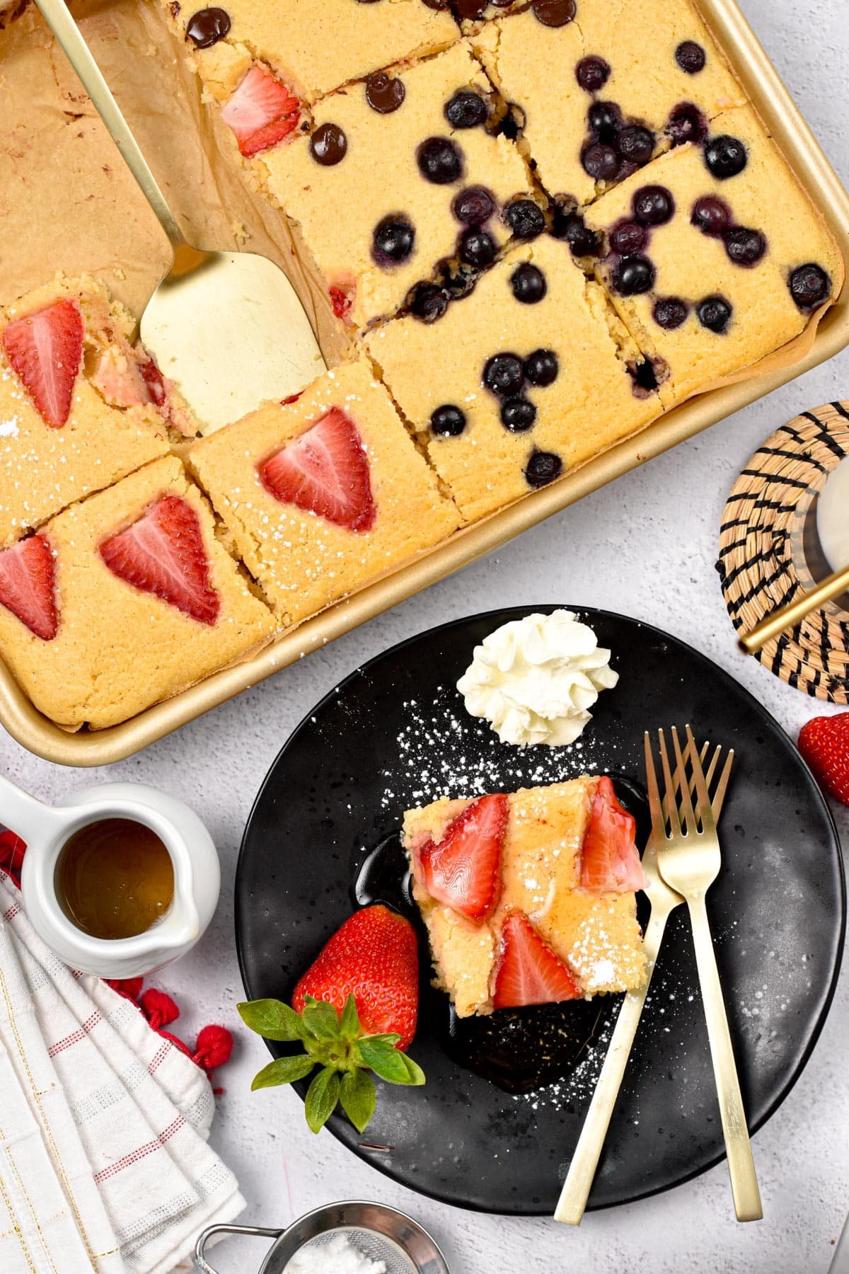 Vegan sheet pan pancakes decorated with strawberries and blueberries next to a plate with a square served with vegan whipped cream.