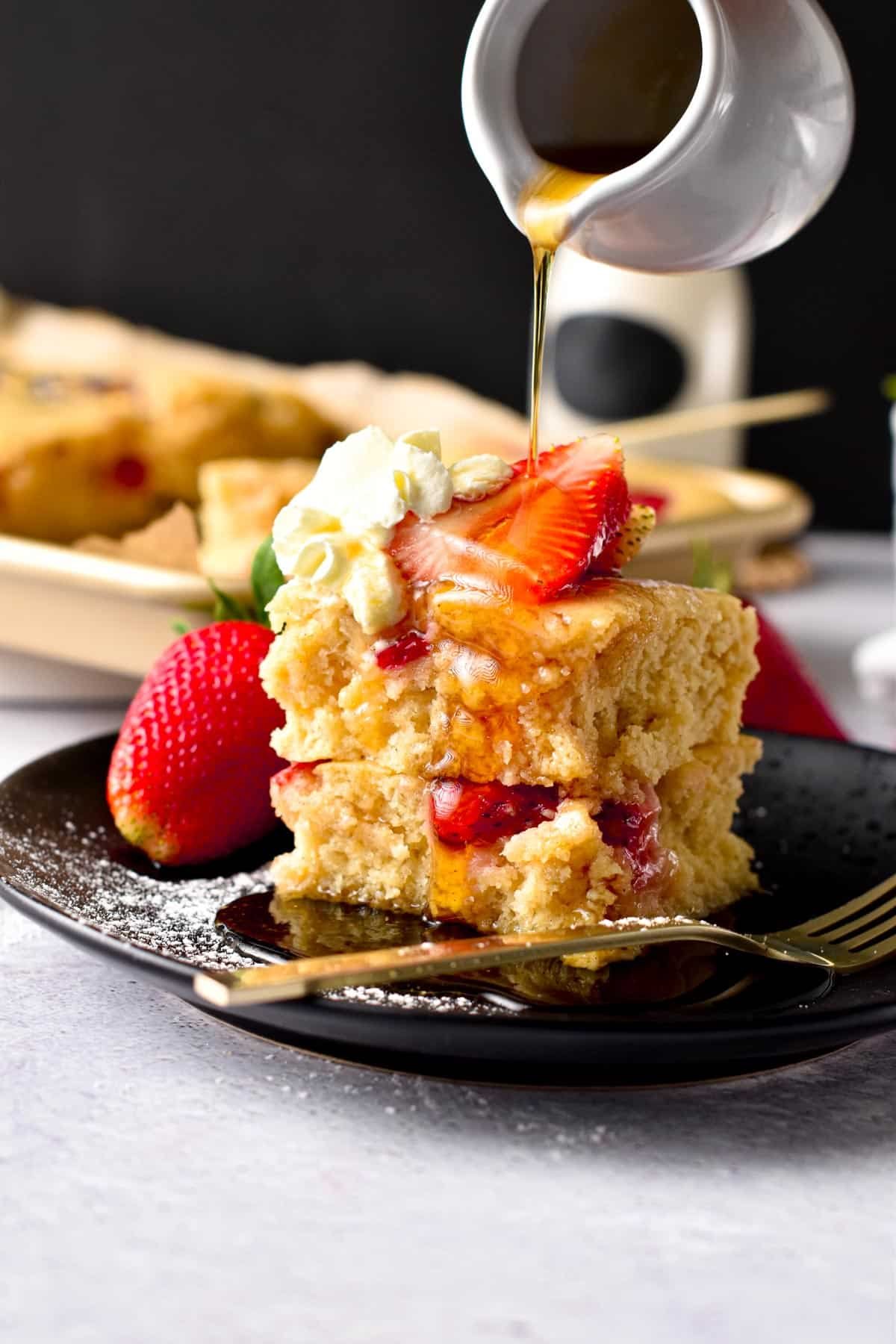 A plate filled with a stack of two vegan sheet pan pancakes, fresh strawberries, dairy-free whipped cream and a drizzle of maple syrup