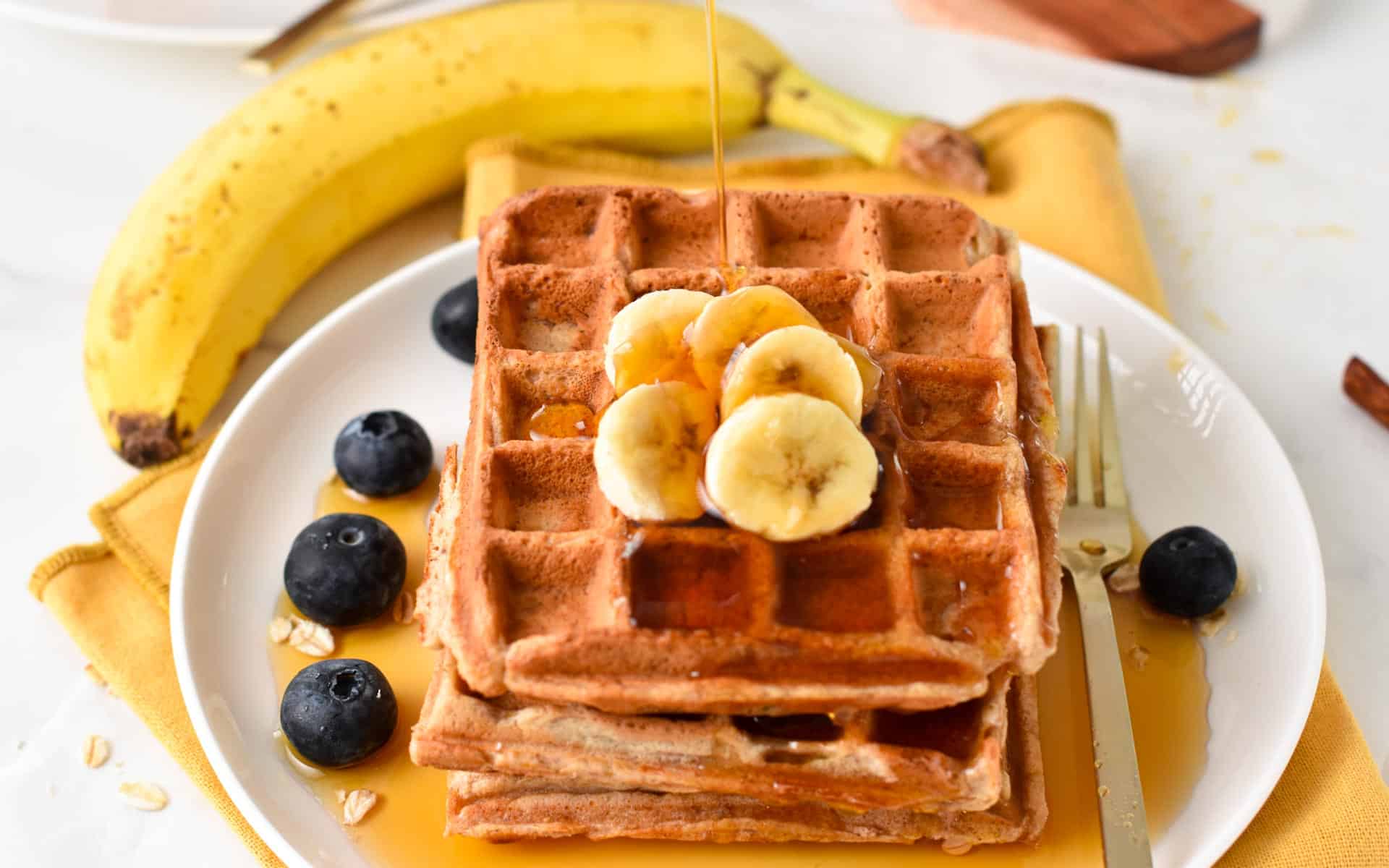a stack of Healthy Banana Waffles with banana slices on top, blueberries around in the plate and a drizzle of syrup on top