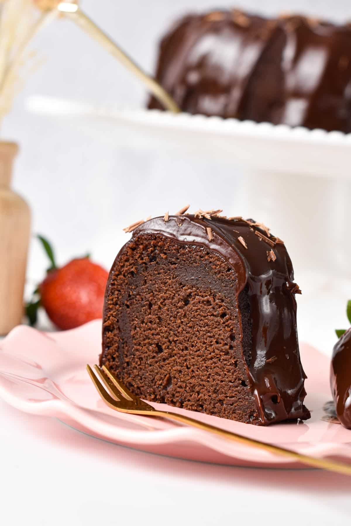 A slice of vegan chocolate Bundt cake on a pink plate with a golden fork on side and strawberries in the background.