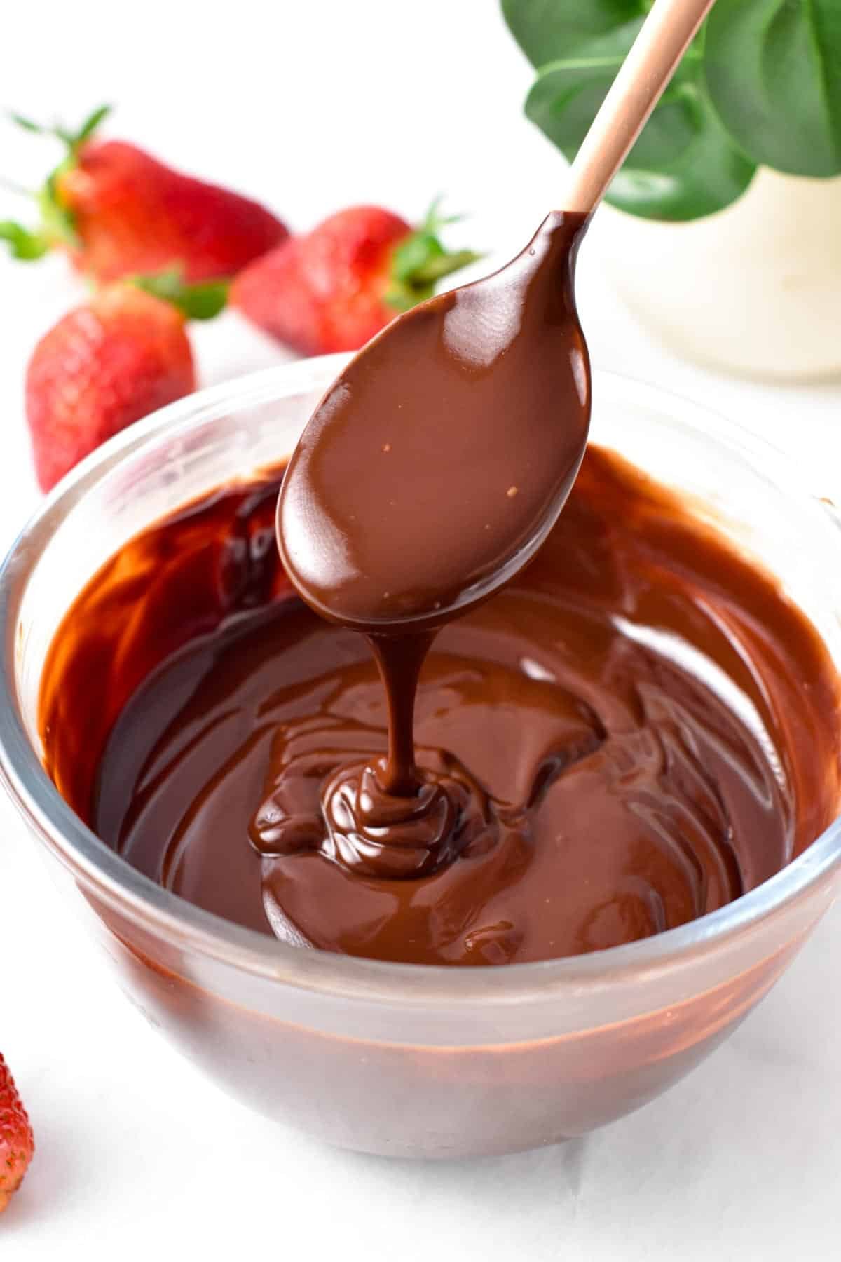 A spoon drizzling chocolate ganache in a glass bowl filled with a large batch of vegan chocolate ganache and strawberries in the background.