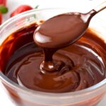 a spoon drizzling chocolate ganache in a glass bowl filled with a large batch of vegan chocolate ganache