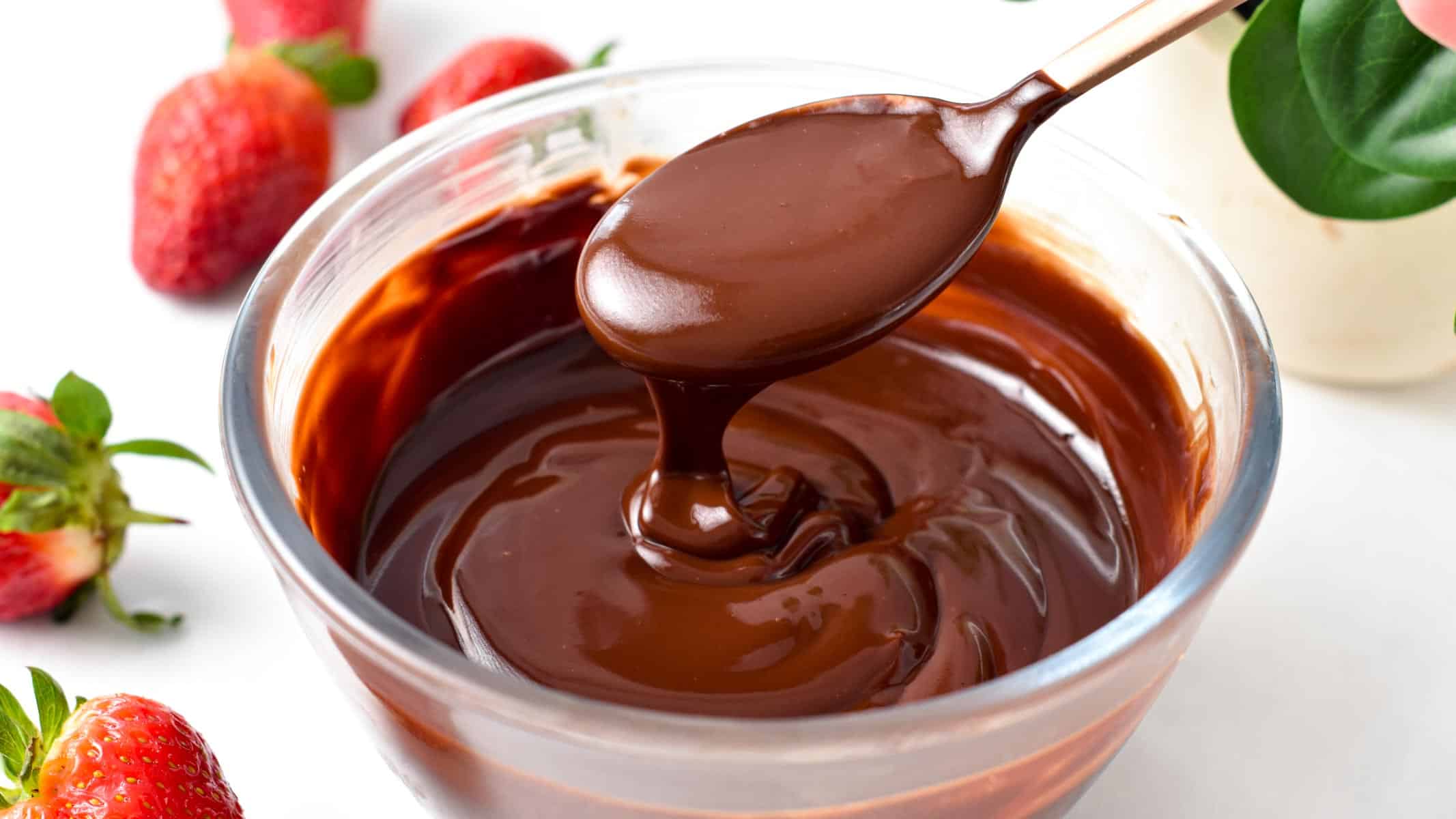 a spoon drizzling chocolate ganache in a glass bowl filled with a large batch of vegan chocolate ganache