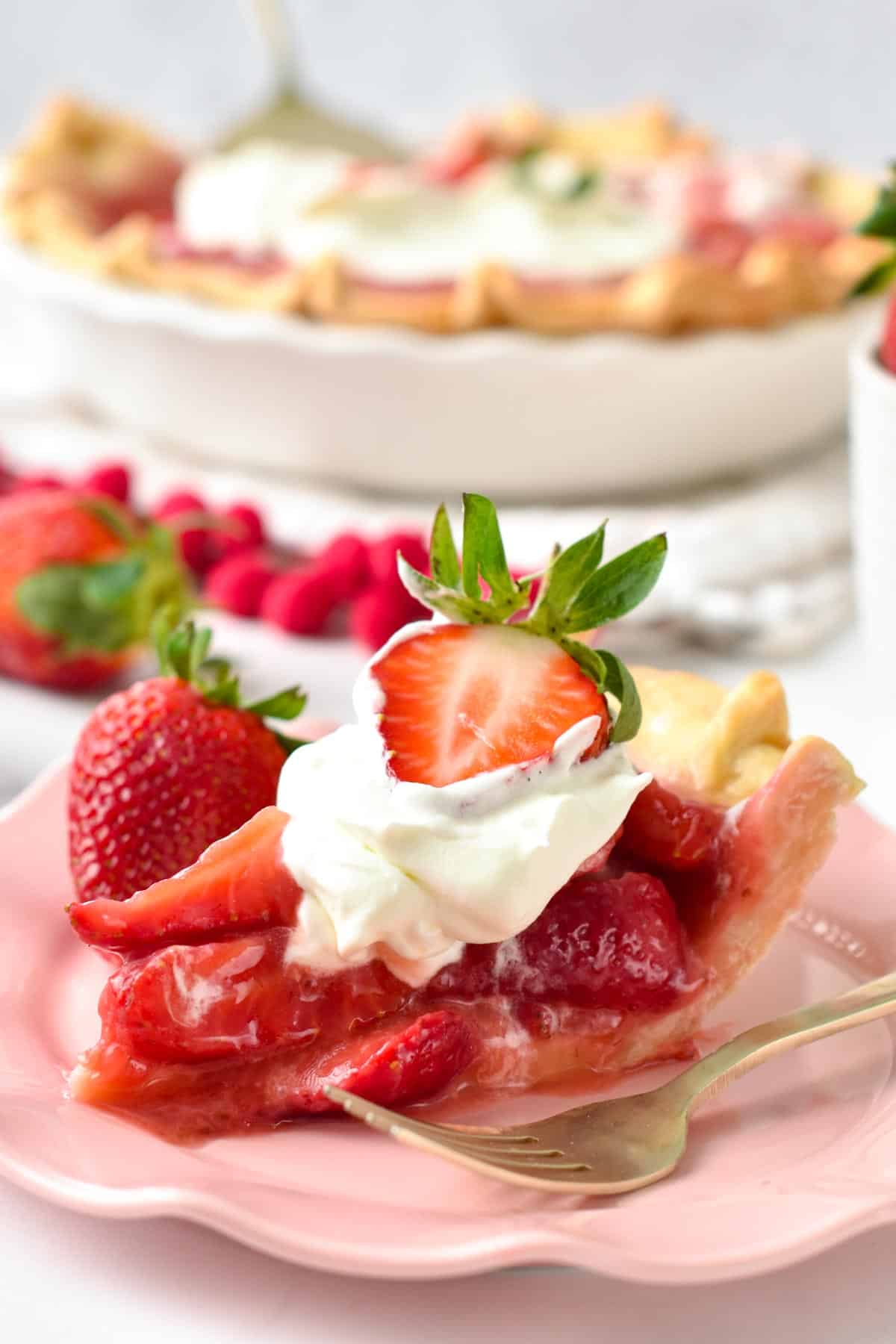 A slice of Vegan Strawberry Pie topped with dairy-free whipped cream, a slice a strawberry and the whole strawberry pie in the background