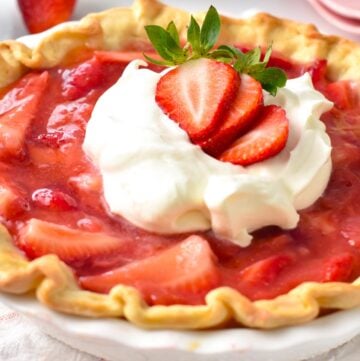 a vegan strawberry pie in a white pie dish topped with dairy-free whipped cream, and fresh strawberry slices