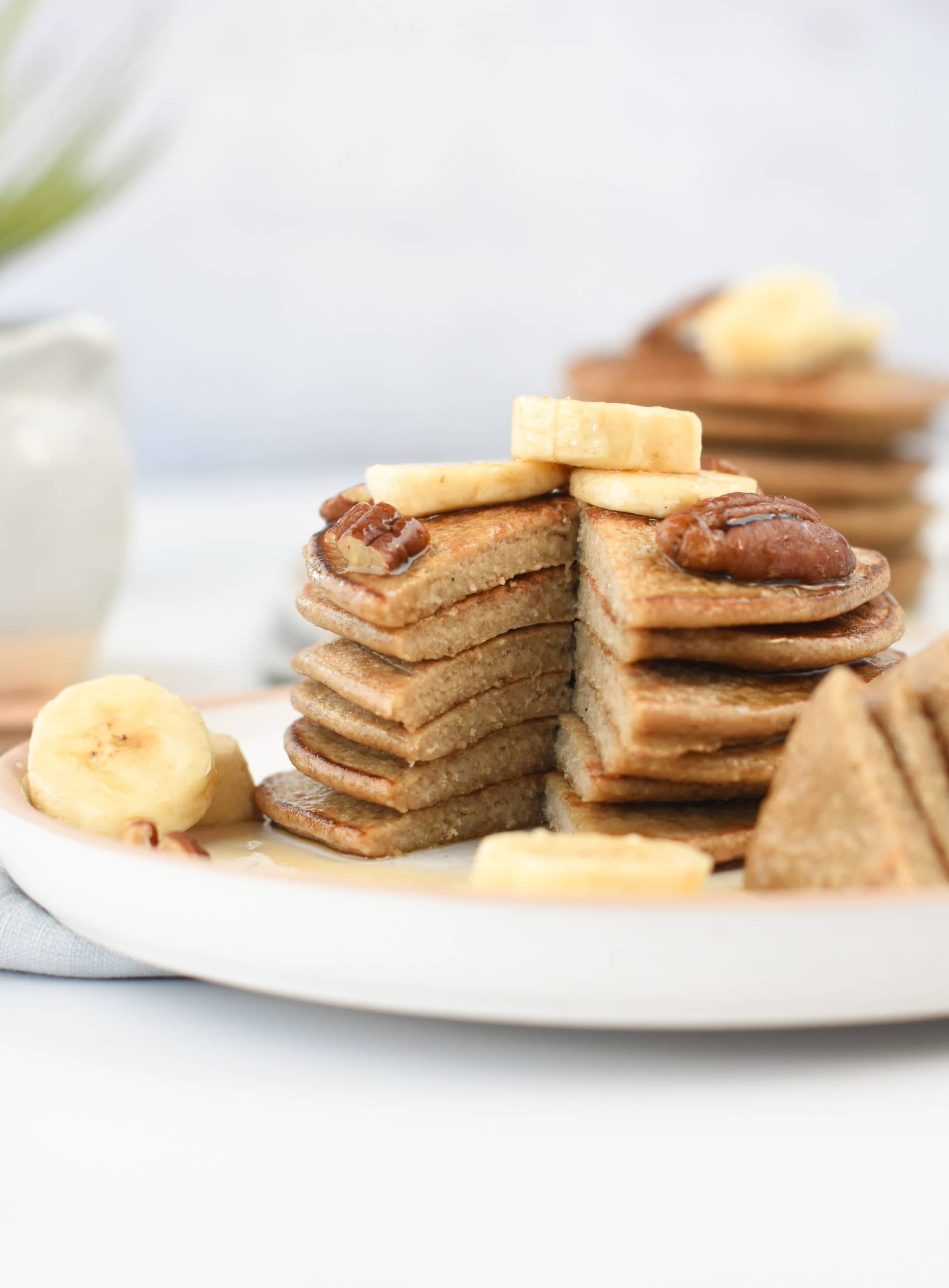 3-Ingredient Banana Oat Pancakes served as a stack on a white plate and decorated with banana slices, pecan nuts, and maple syrup.