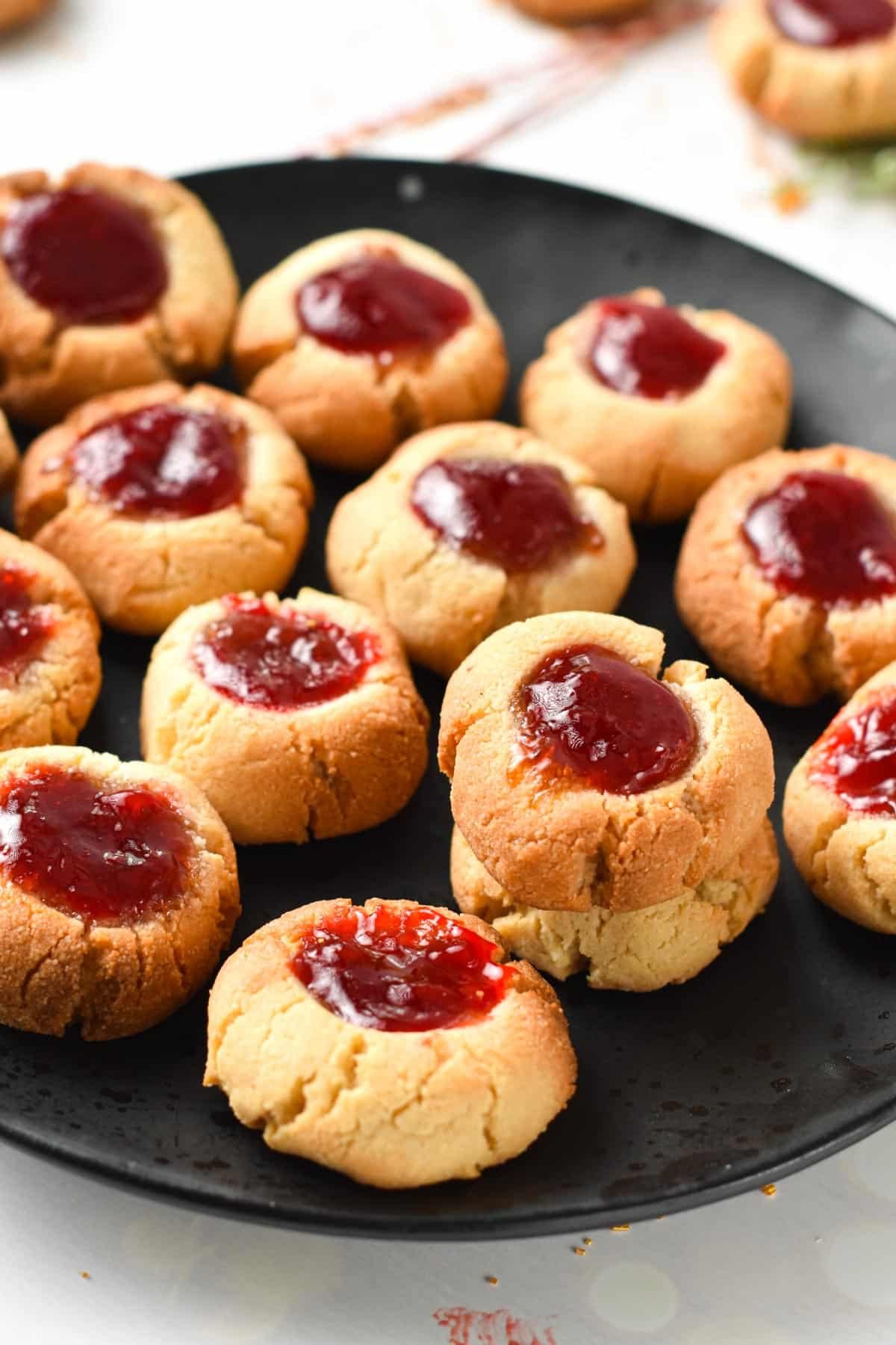 A black plate filled with almond flour thumbprint cookies filled with vibrant red jam.