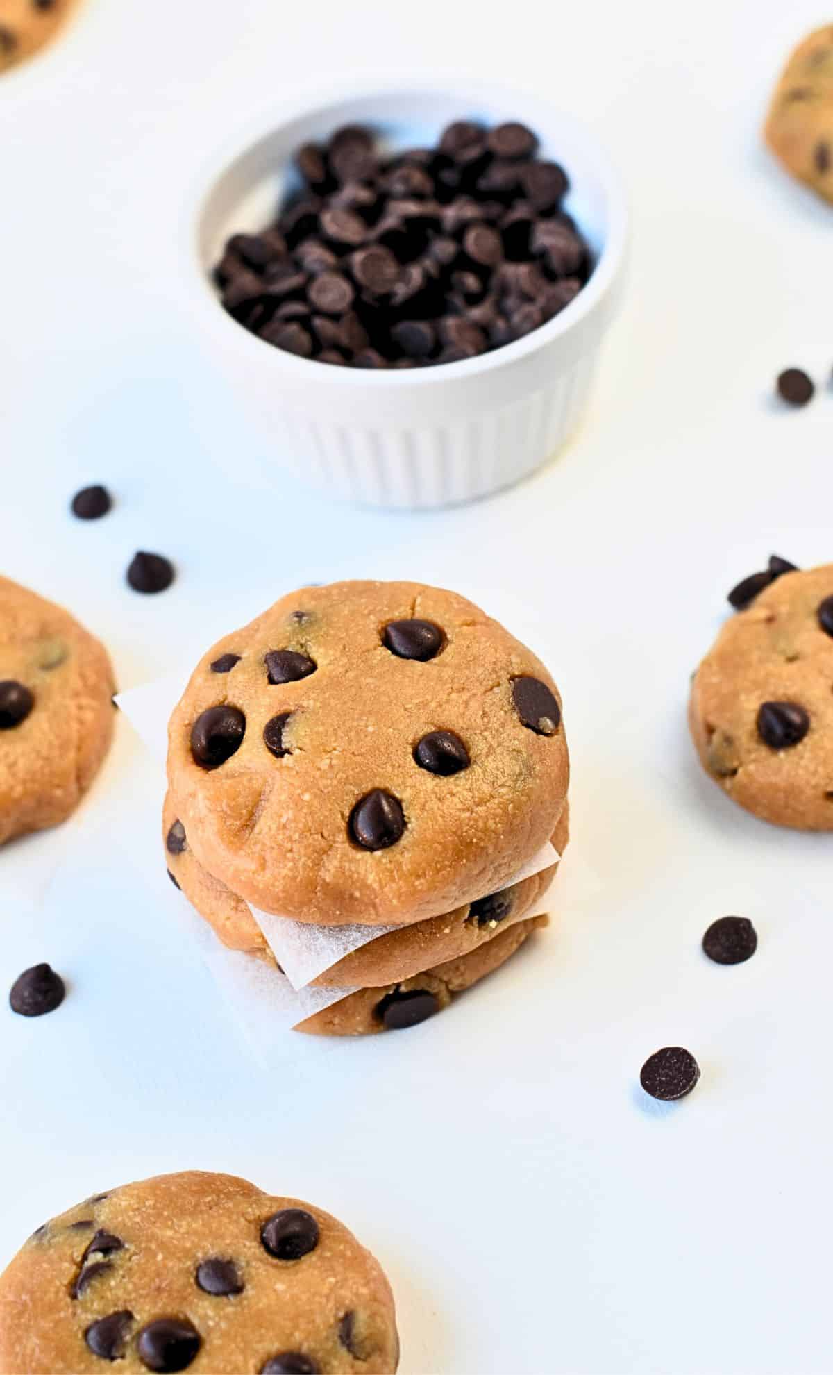 No-Bake Chocolate Chip Cookies stacked on a white table in front of a bowl with chocolate chips.