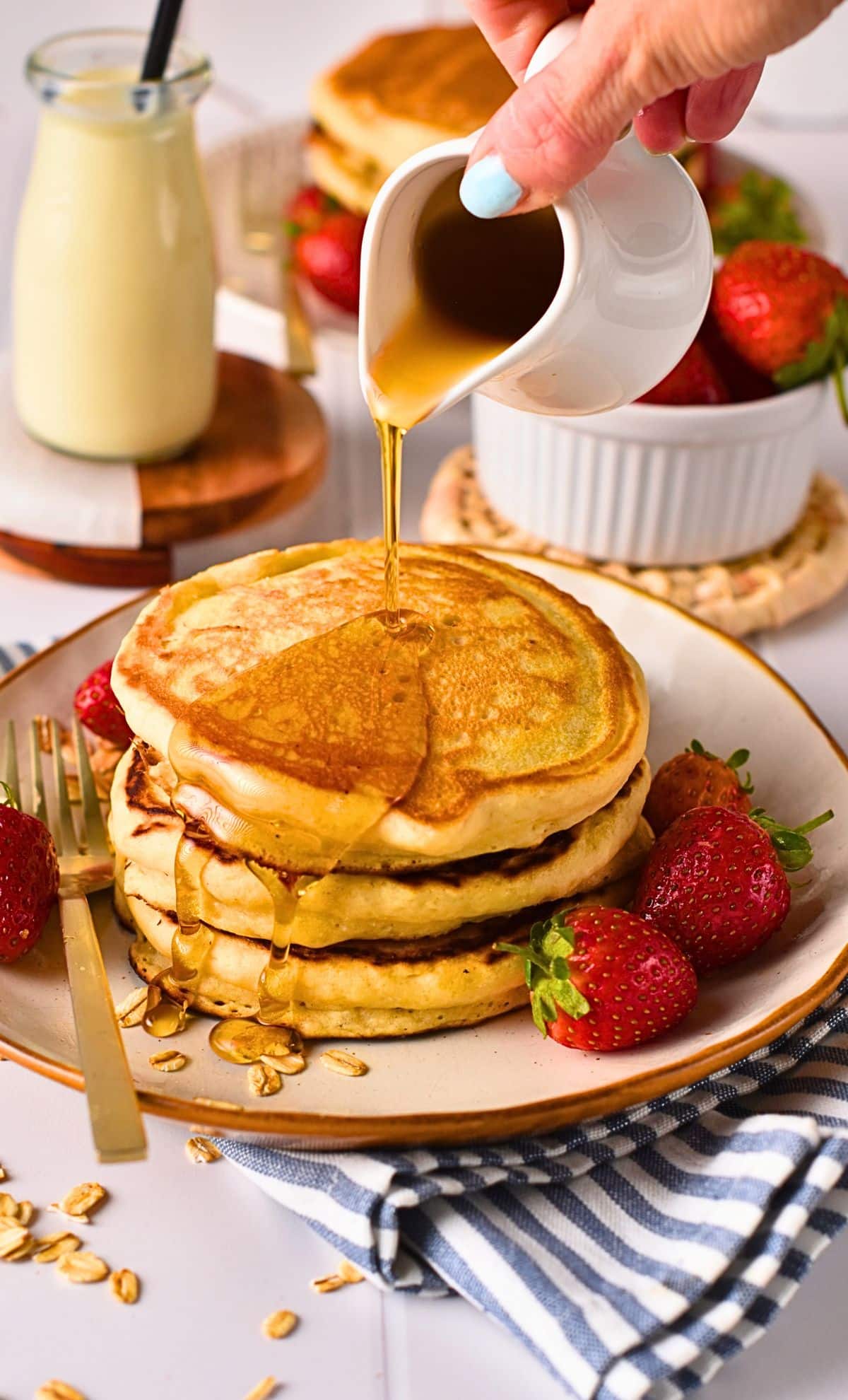 Pouring maple syrup on a stack of vegan pancakes decorated with strawberries on a plate with a golden fork.