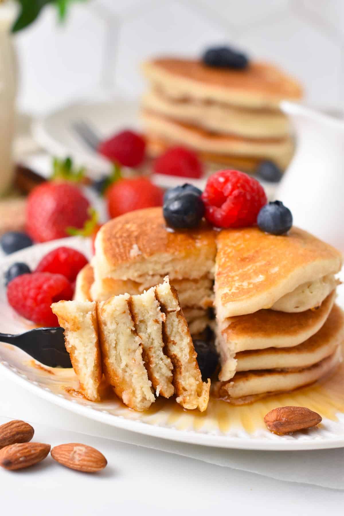 A stack of vegan almond flour pancakes with blueberries and raspberry on top and a fork showing the fluffy texture inside the pancakes.