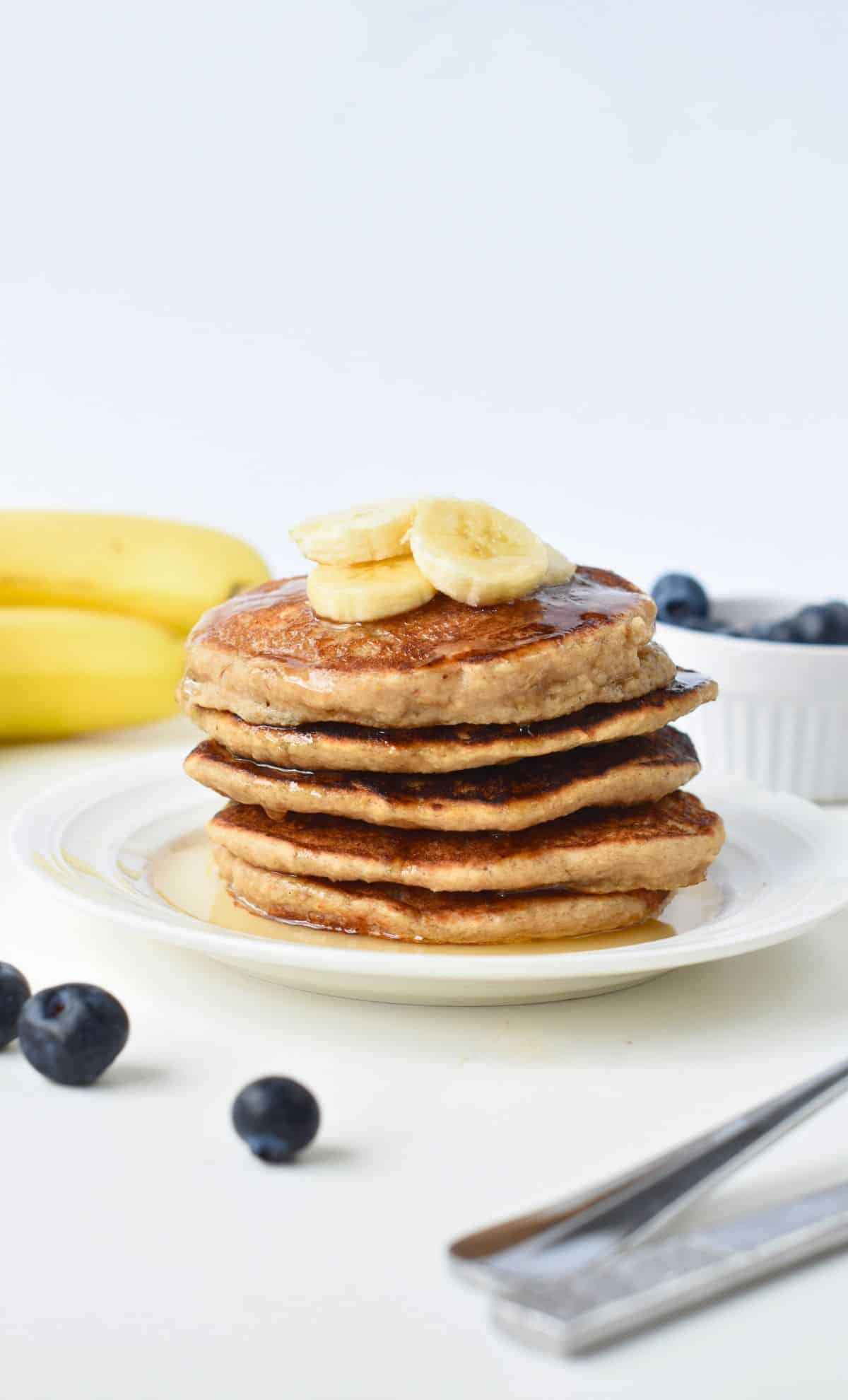 Vegan Banana Oat Pancakes stacked on a white plate, decorated with banana  and maple syrup.