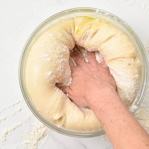 a hand pushing down raised beignet dough in a  glass mixing bowl.