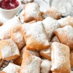 a cooling rack loaded with vegan beignets dusted with powdered sugar and a pot of red jam in the background