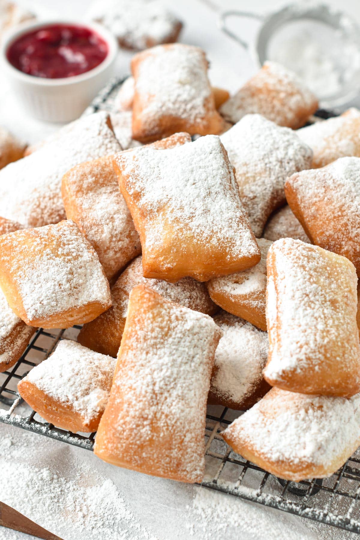 A cooling rack loaded with vegan beignets dusted with powdered sugar and a pot of red jam in the background.