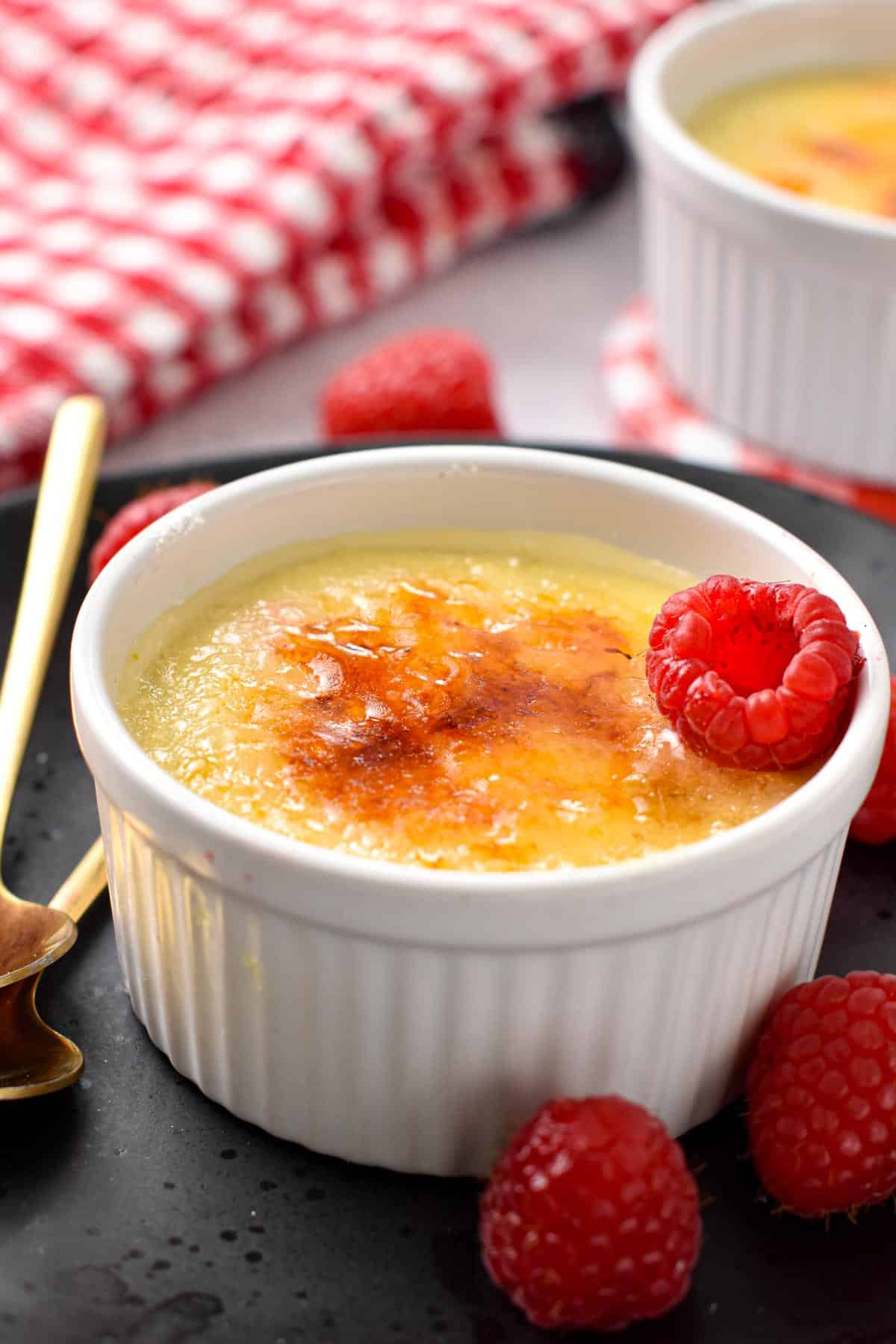 A ramekin filled with a creme brulee mixture topped with crunchy caramel golden layer and a fresh raspberry on the side.