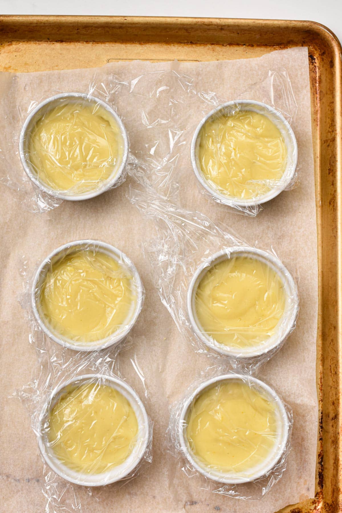 6 creme brulee cooling down in their white ramekin and covered with a piece of plastic wrap