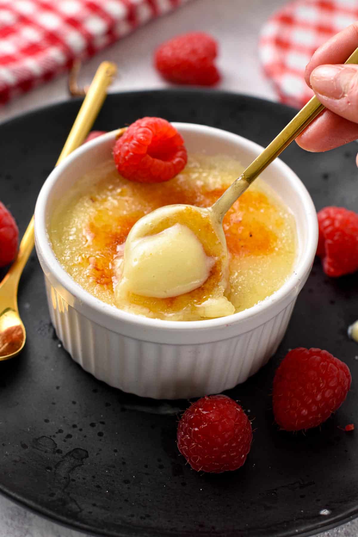 A golden spoon digging in a creamy creme brulee with crispy caramel topping and a raspberry on side.