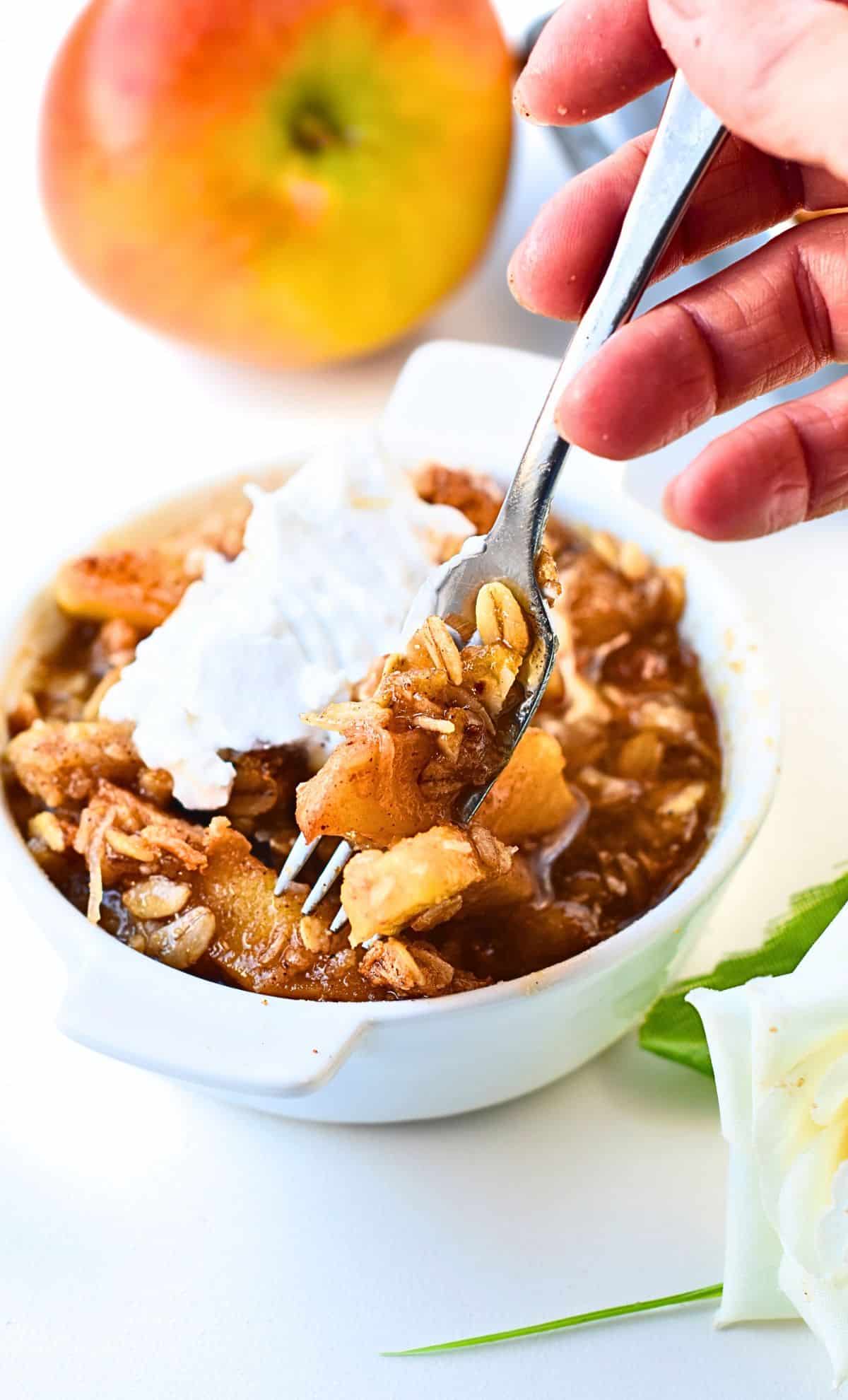 A fork taking some vegan gluten-free apple crisp off a ramekin decorated with vegan whipped cream.