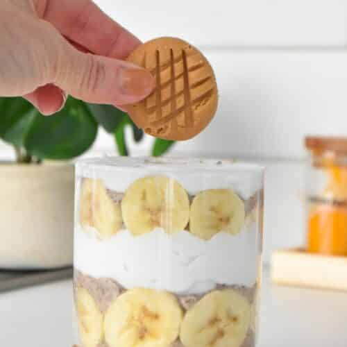 a jar filled with layers of banana chia seed pudding, coconut yogurt and a hand holding a no bake peanut butter cookie on top.