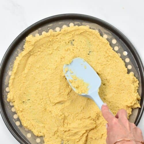 A blue silicone spatula spreading chickpea pizza dough on a pizza rack.