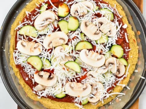 Cooked chickpea pizza crust with marinara sauce, sliced mushrooms, zucchinis and vegan cheese, on a round pizza baking sheet.
