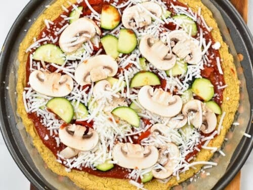 Cooked chickpea pizza crust with marinara sauce, sliced mushrooms, zucchini and vegan cheese, on a round pizza baking sheet.