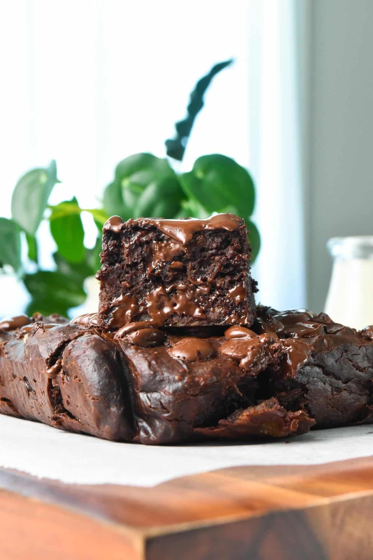 A piece of healthy banana brownies stacked on top of a brownie and a green plant in the back.